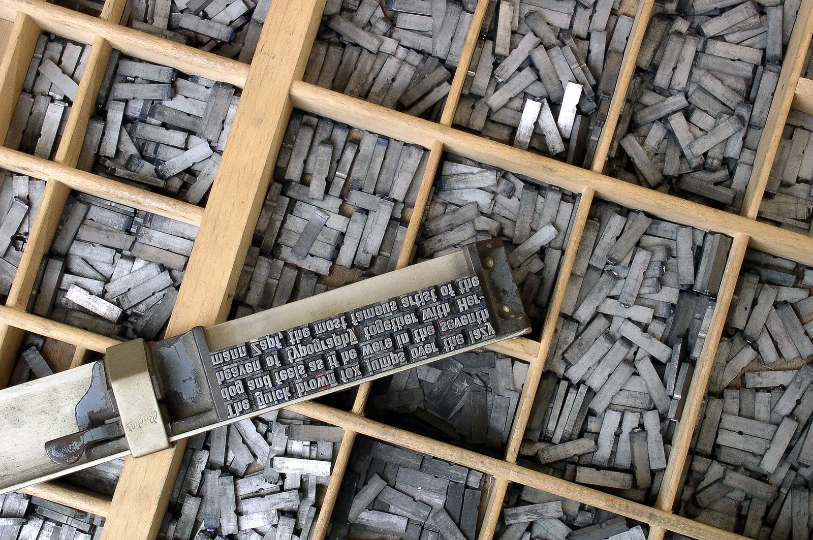 A wooden type case full of grey metal letter blocks, with a composing stick holding a few lines of type set in mirror image across the middle
