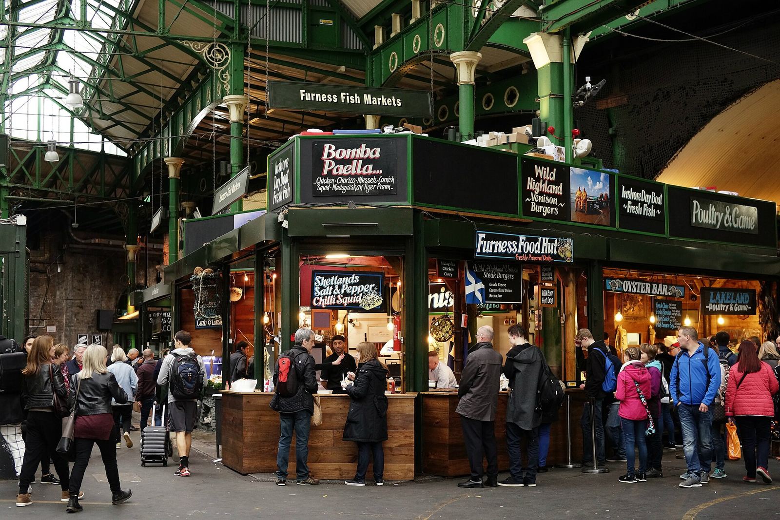 Food stalls under the green iron roof of Borough Market, with signs for paella and oysters and a lunchtime crowd queuing at the counters