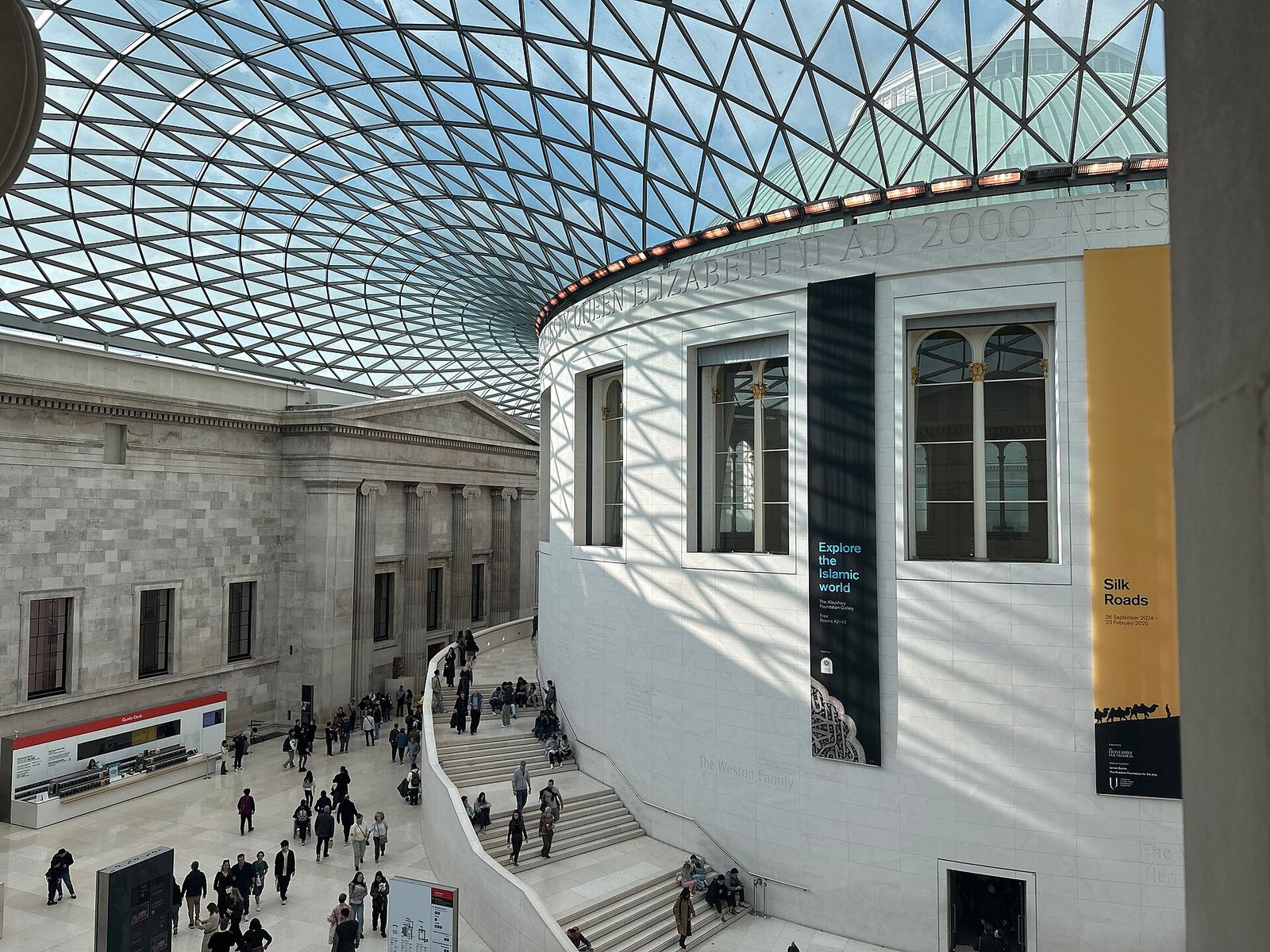 The British Museum's Great Court: a glass-and-steel lattice roof curving over the white drum of the old Reading Room, with visitors on the curving staircase and the courtyard floor