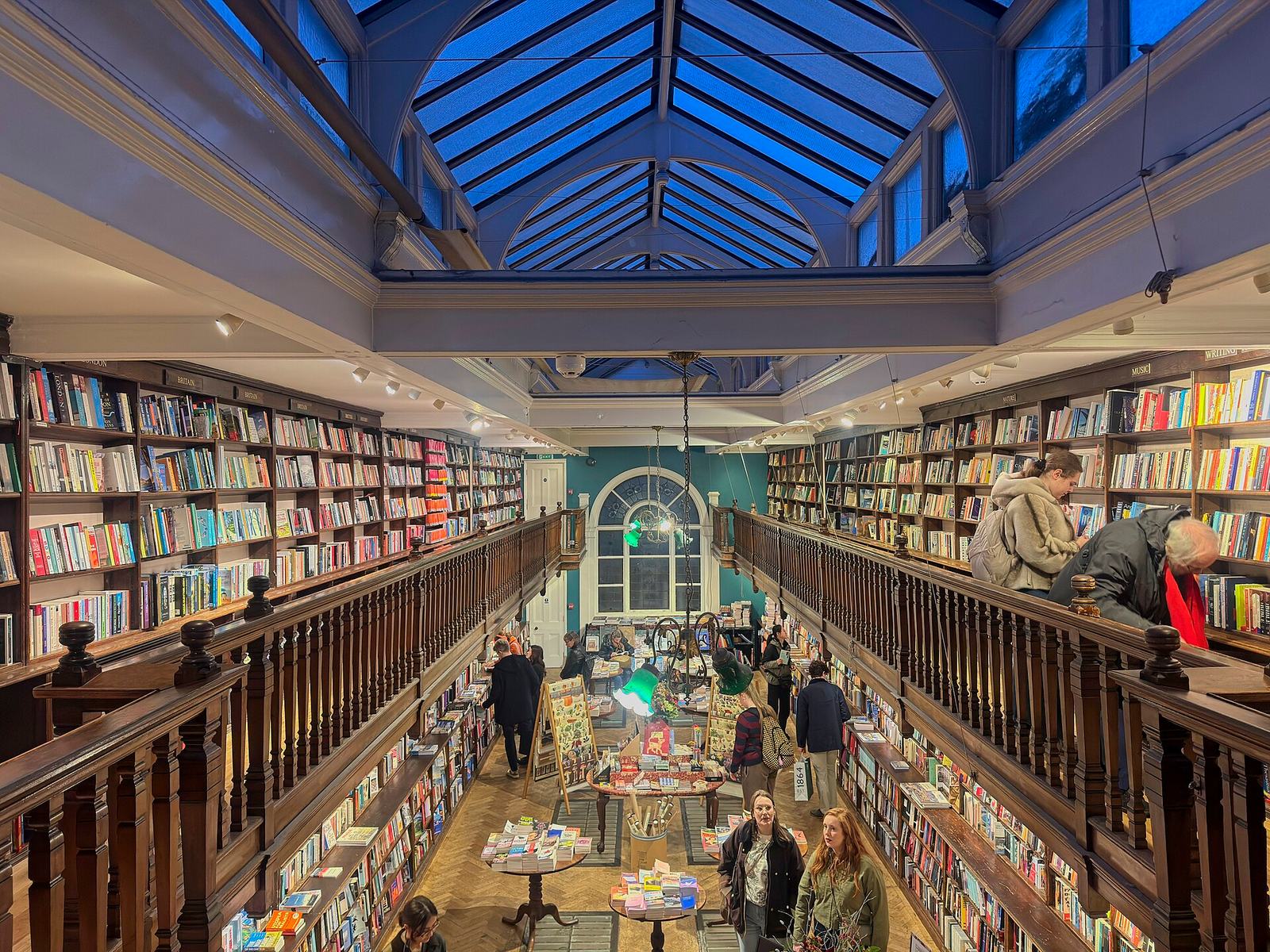 Inside Daunt Books in Marylebone: long oak galleries of bookshelves on two levels under a glass skylight, leading to an arched stained-glass window, with tables of books below