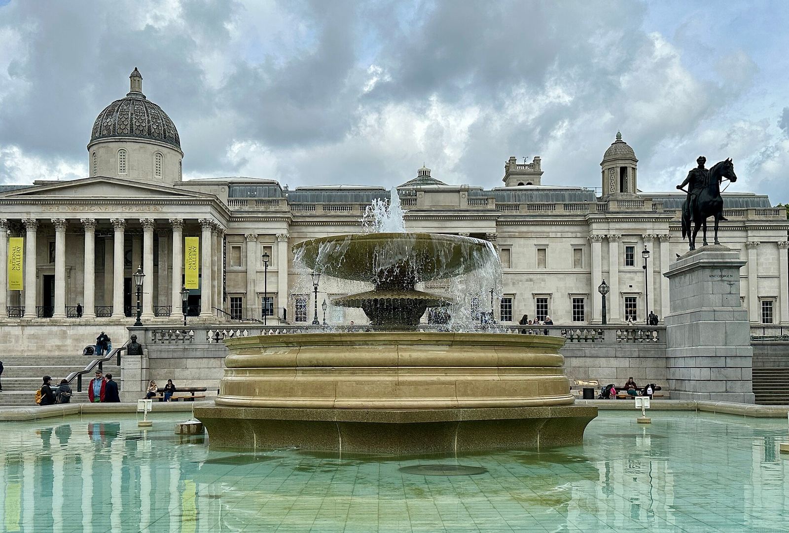 The columned front and dome of the National Gallery behind one of the Trafalgar Square fountains, under a cloudy sky
