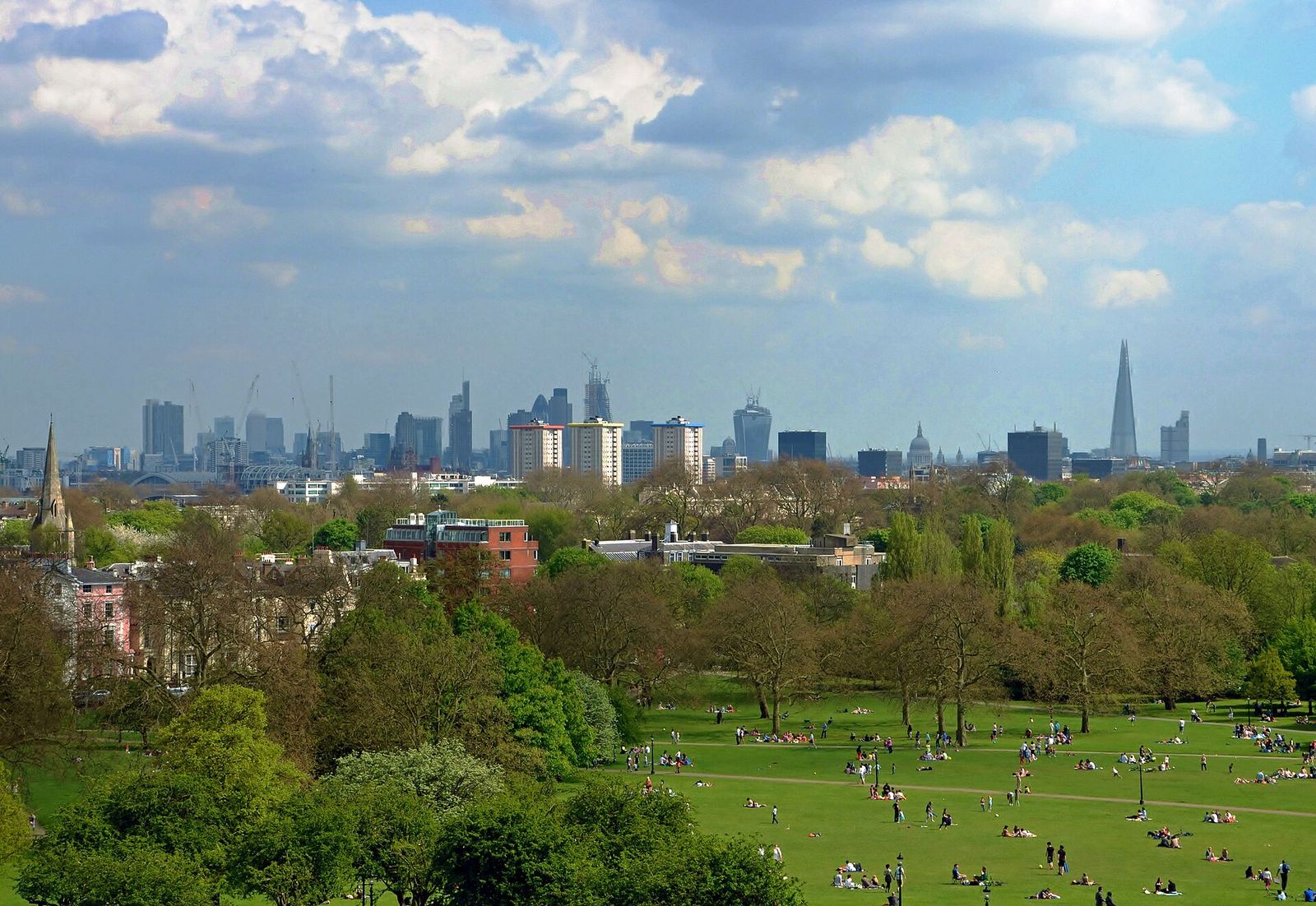 Central London from Primrose Hill: people on the grass in the park below, trees and rooftops, and the skyline of the City and the Shard on the horizon under white clouds