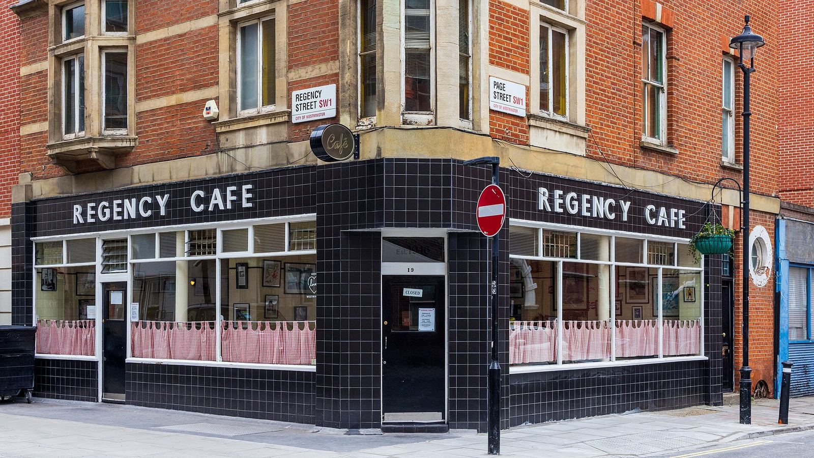 The Regency Café on a Westminster street corner: a black-tiled ground floor with REGENCY CAFE in white letters over both frontages and pink gingham half-curtains in the windows