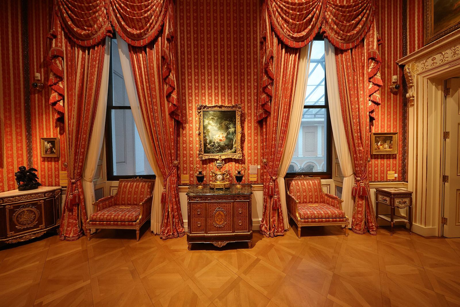A red-silk room at the Wallace Collection with Fragonard's The Swing in a gilt frame between two tall curtained windows, above an inlaid cabinet and two armchairs
