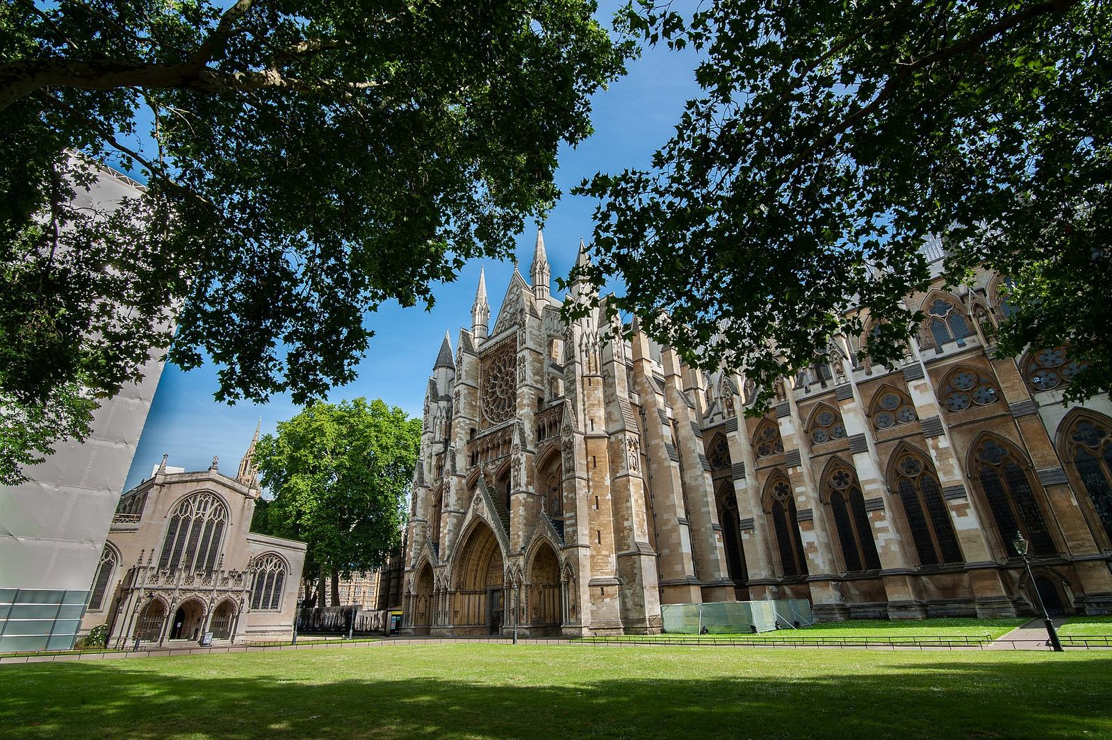 The north front of Westminster Abbey, with its Gothic spires and rose window, seen across a green lawn under trees, with St Margaret's Church to the left