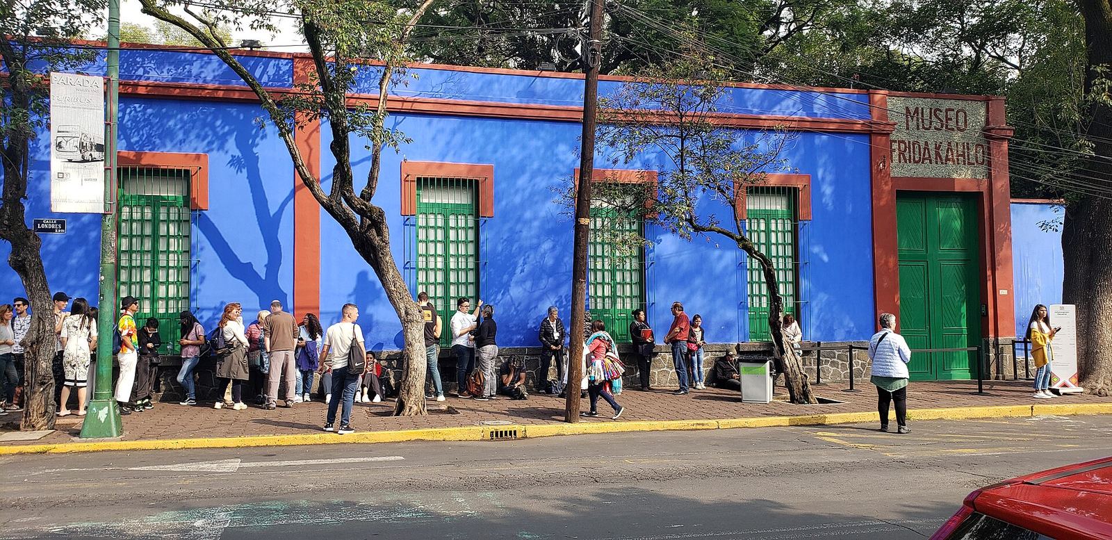 The cobalt-blue outer walls of the Casa Azul on Calle Londres, with green shuttered windows, red trim and a line of visitors waiting under the trees