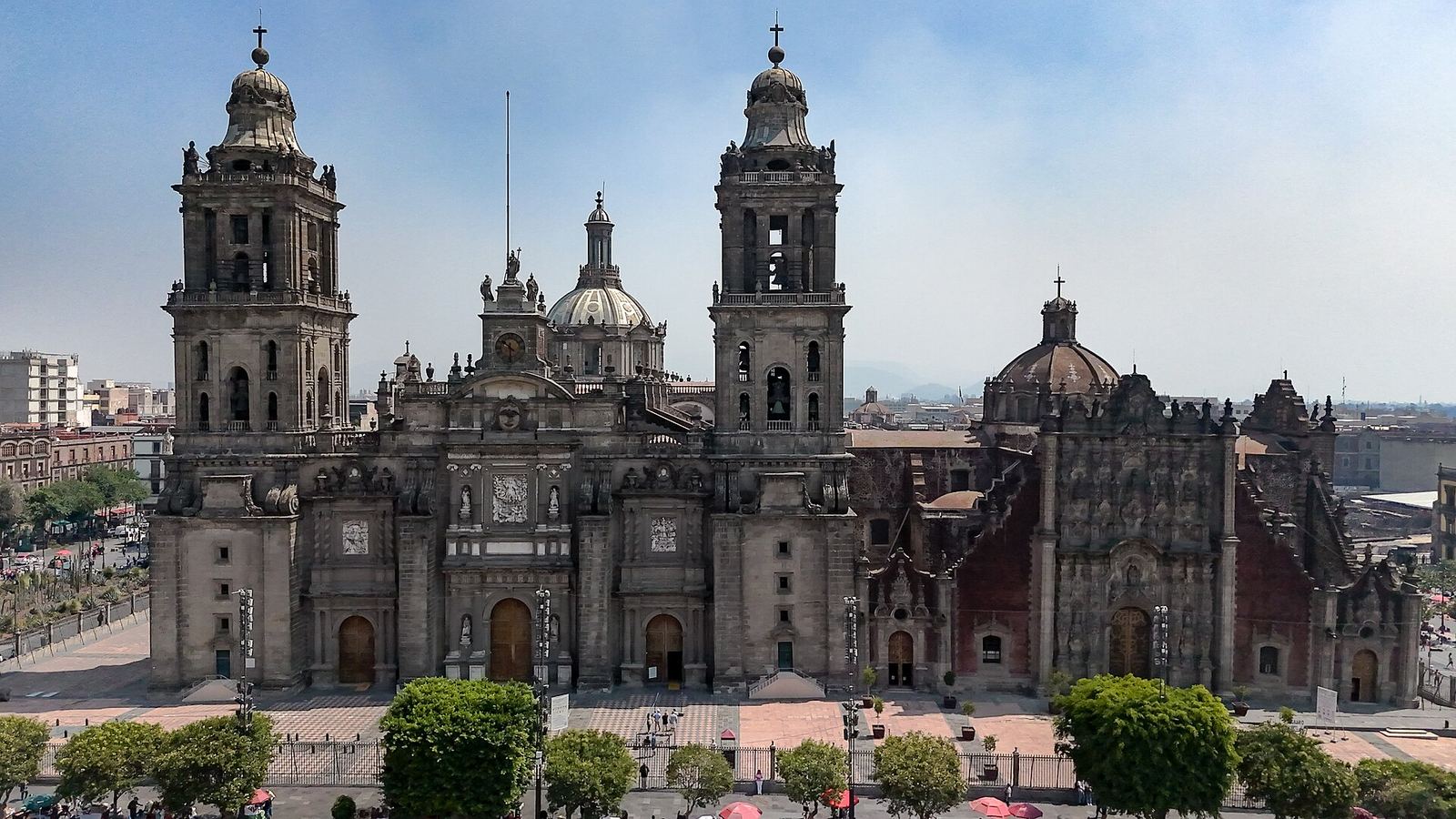 The Metropolitan Cathedral of Mexico City seen from across the Zócalo: a wide baroque facade with two tall bell towers and domes, trees and a low fence in front, under a pale blue sky