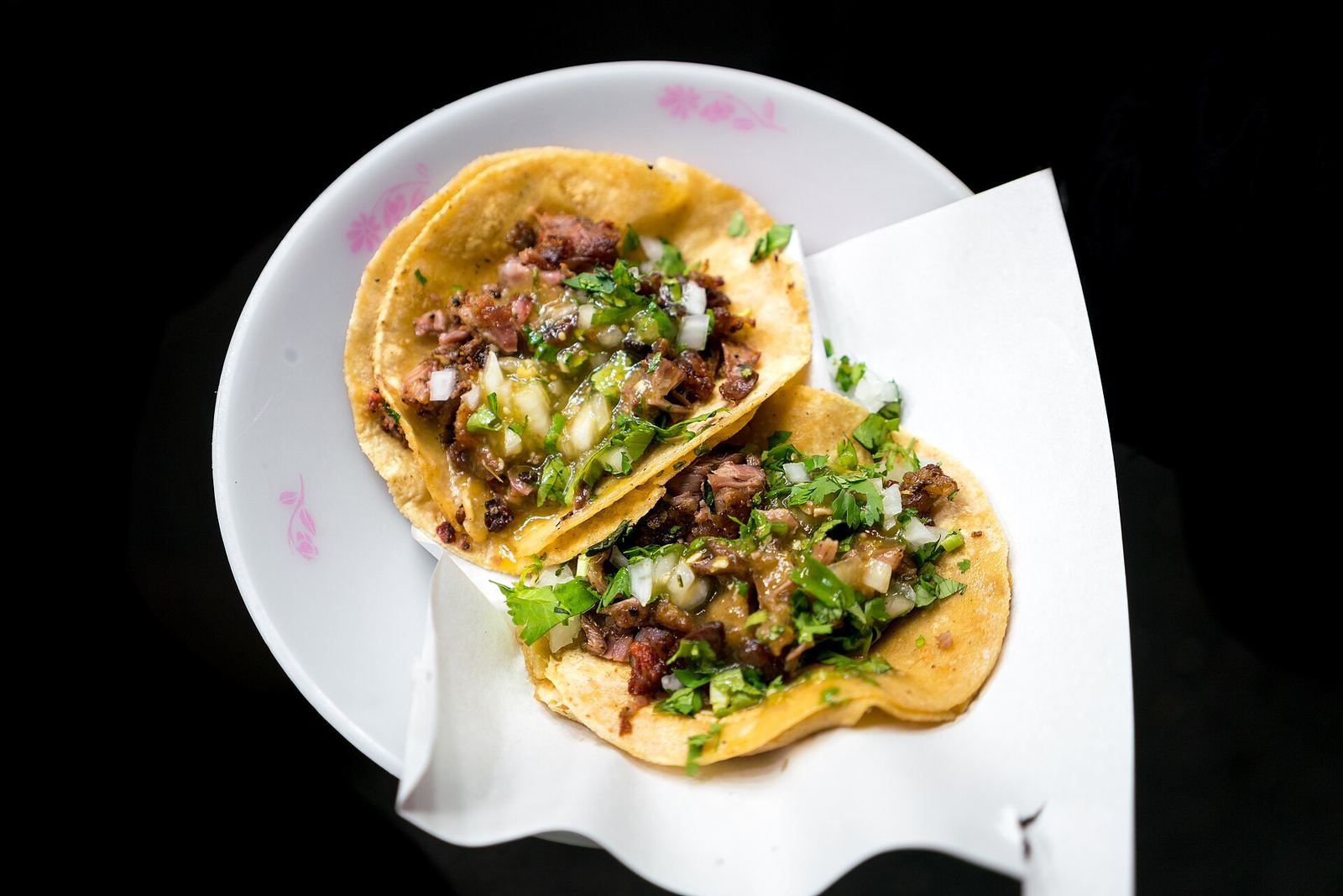 Two tacos from Los Cocuyos on a white plate and paper, topped with chopped onion and cilantro, on a black background