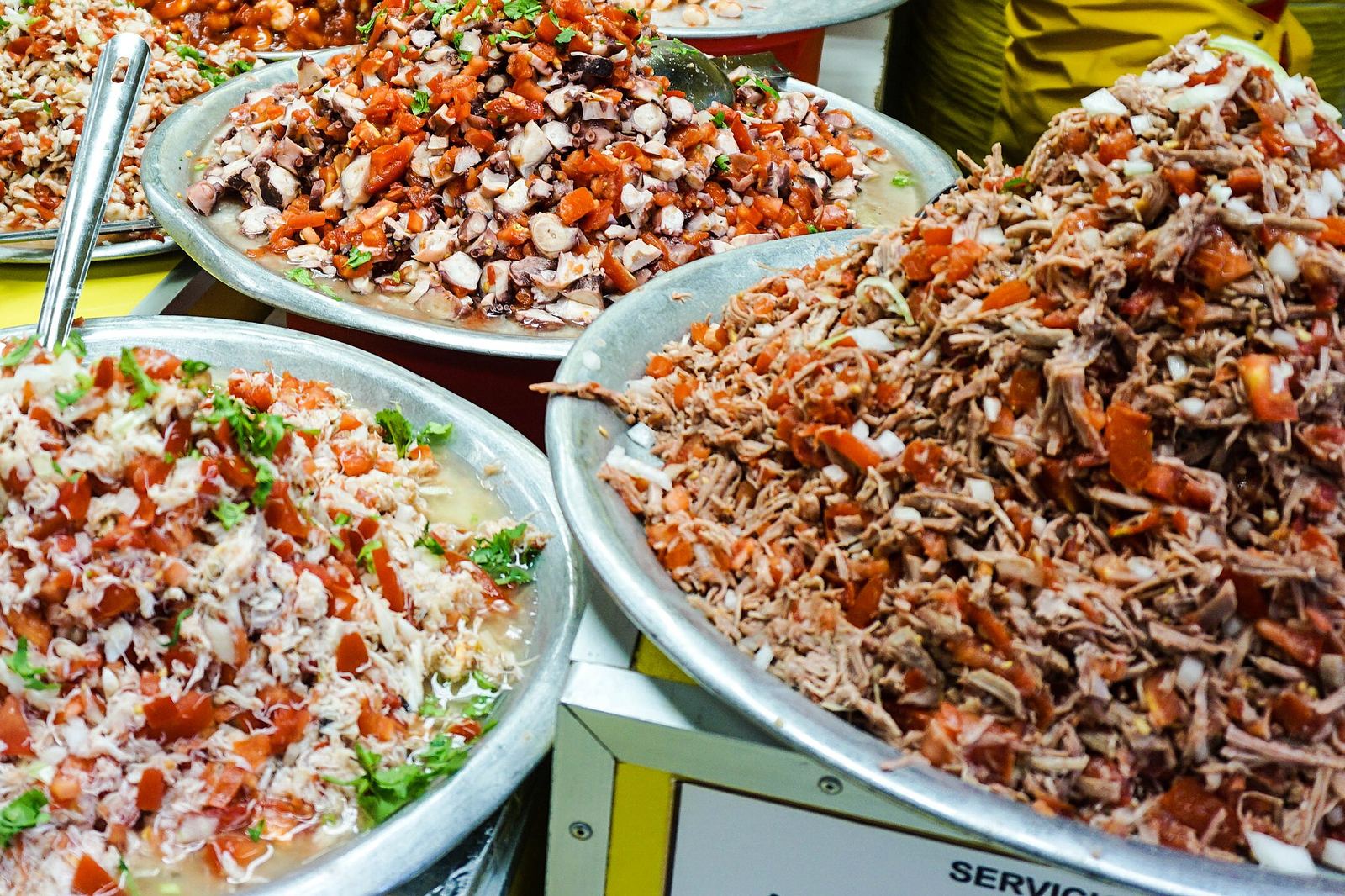 Big metal bowls of tostada toppings at a Mercado de Coyoacán stall: chopped octopus with tomato, crab with cilantro and shredded meat with onion