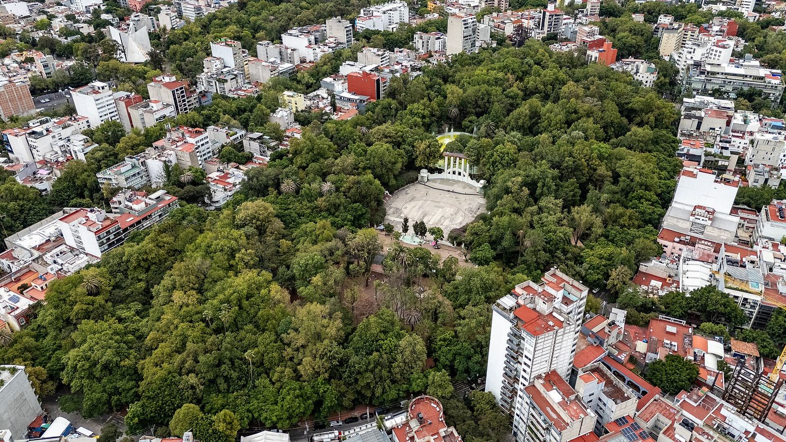 Aerial view of Parque México in Condesa: a dense green canopy of trees ringed by apartment buildings, with the park's open-air forum in a clearing