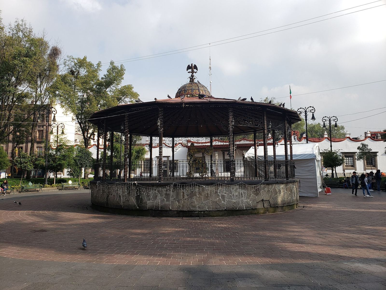 The domed bandstand in Coyoacán's Plaza Hidalgo, an octagonal kiosk topped by an eagle, on an empty paved square framed by trees