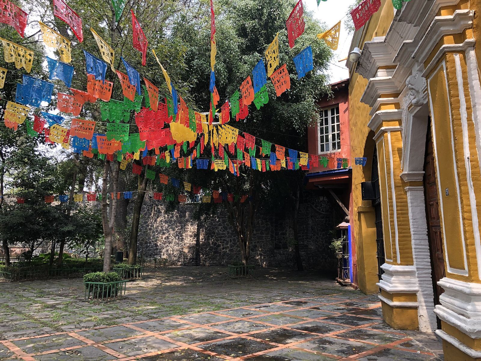 The yellow Capilla de Santa Catarina in Coyoacán, its baroque doorway and walls hung with strings of colourful papel picado over a paved plaza with trees