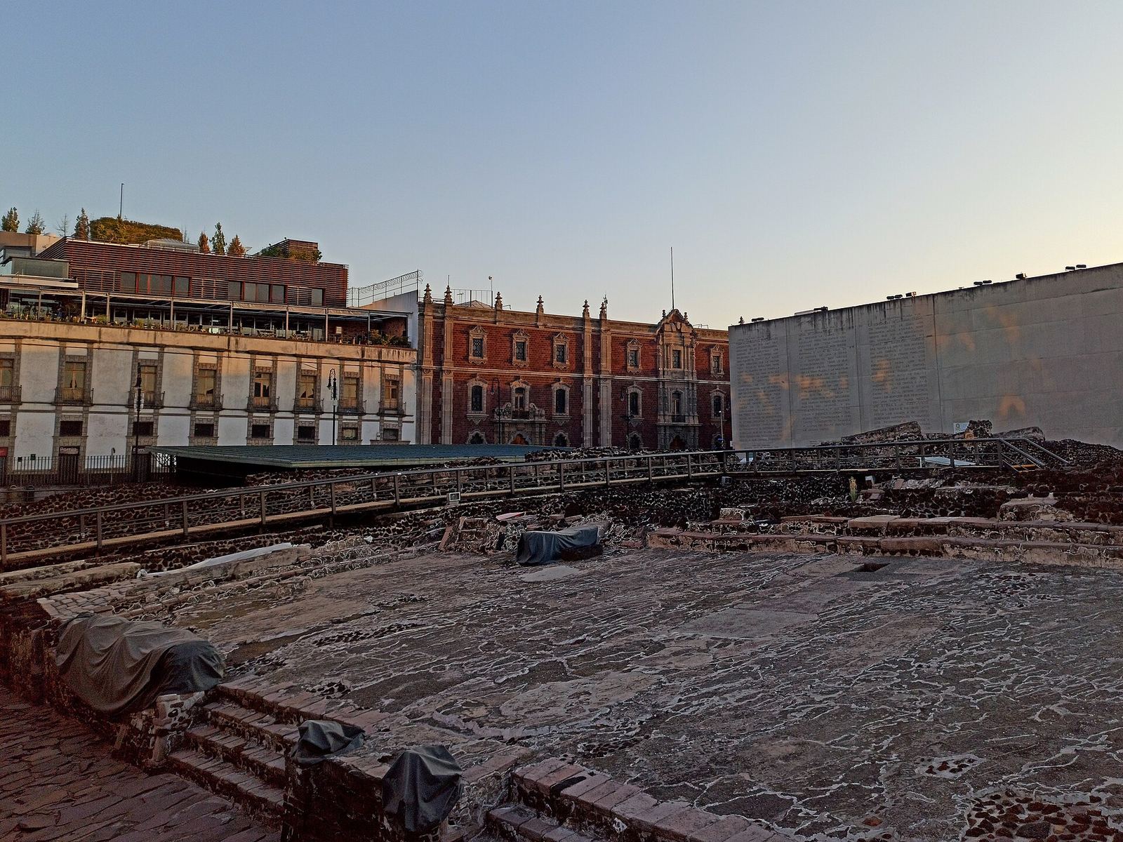 The excavated ruins of the Templo Mayor, with stone walls and stairways in a sunken site, walkways around the edge and colonial buildings of the Centro behind