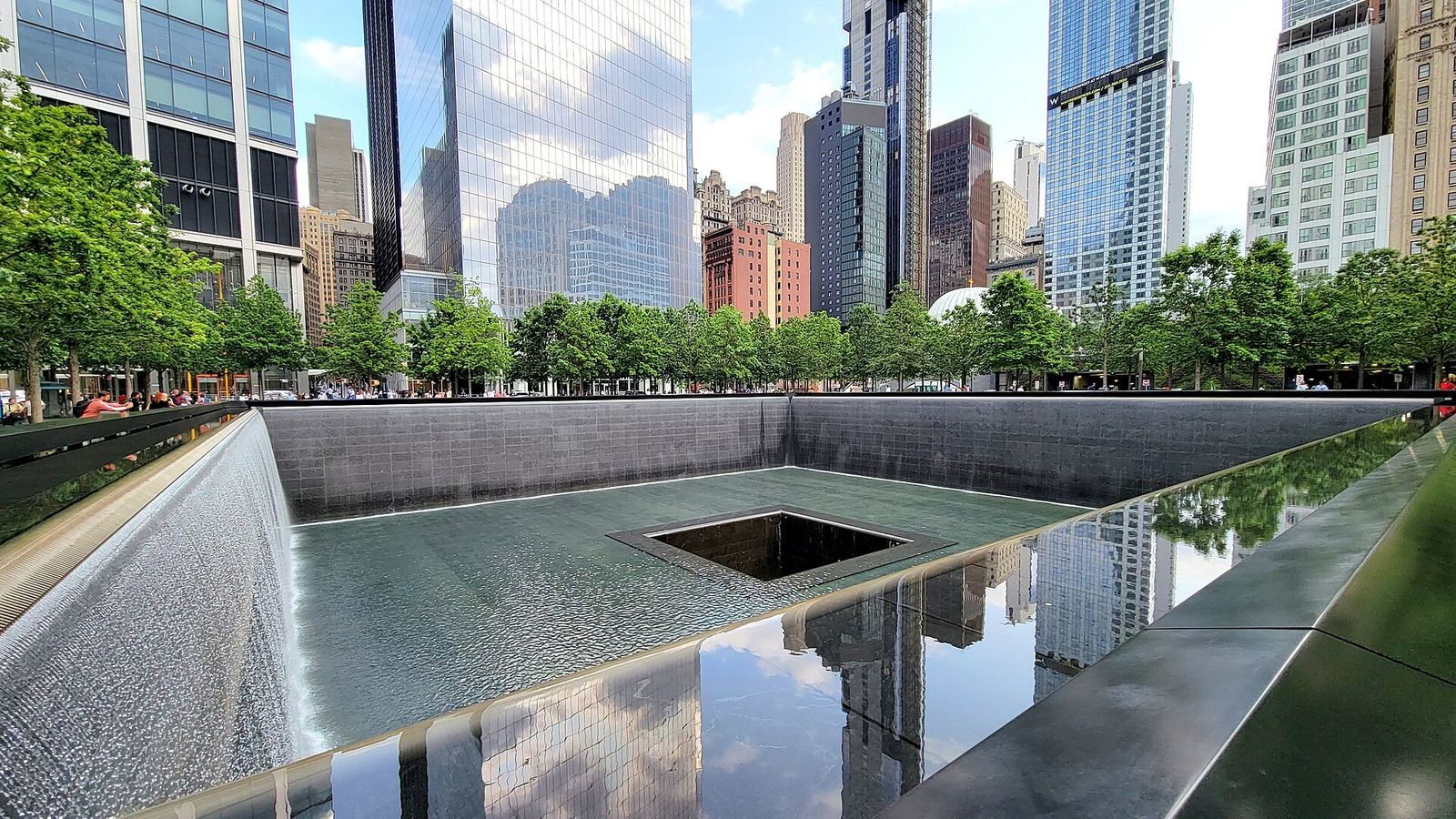 The South Pool of the 9/11 Memorial: water falls down dark granite walls into a square void, with trees and Lower Manhattan's glass towers reflected in the pool