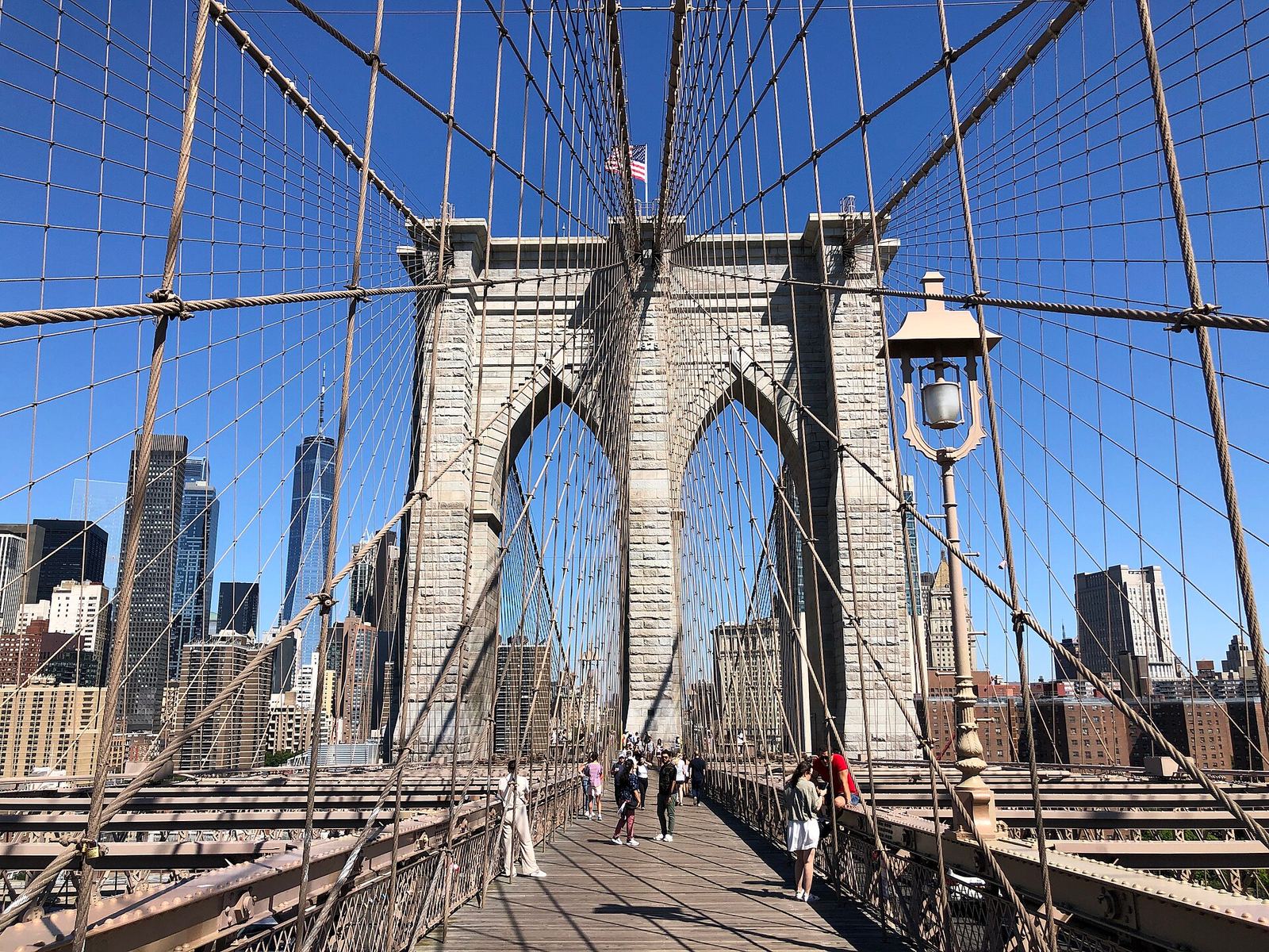 The wooden promenade of the Brooklyn Bridge leading to its stone Manhattan tower with pointed Gothic arches, steel cables fanning overhead and Lower Manhattan's towers behind