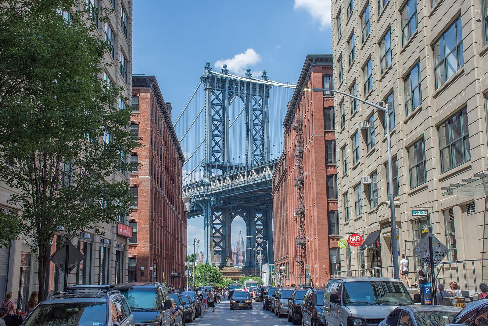 The blue Manhattan Bridge tower framed between red-brick warehouse buildings at the end of Washington Street in DUMBO, with the Empire State Building visible through the tower's arch
