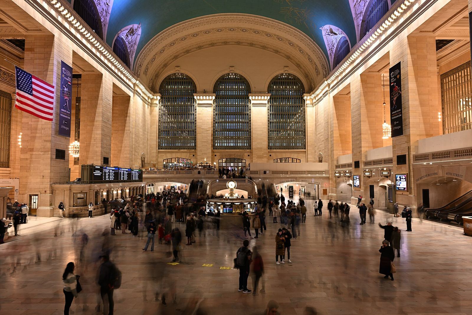 Grand Central Terminal's Main Concourse: tall arched windows at the west end, the blue-green ceiling painted with constellations, an American flag and travelers crossing the marble floor