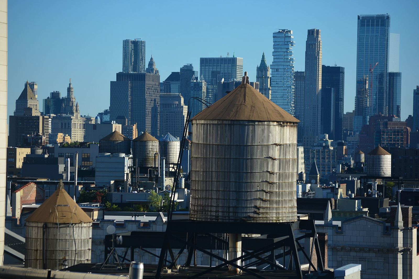 Weathered wooden water tanks with conical roofs on the rooftops of Chelsea, with the skyscrapers of Lower Manhattan rising behind them under a clear blue sky