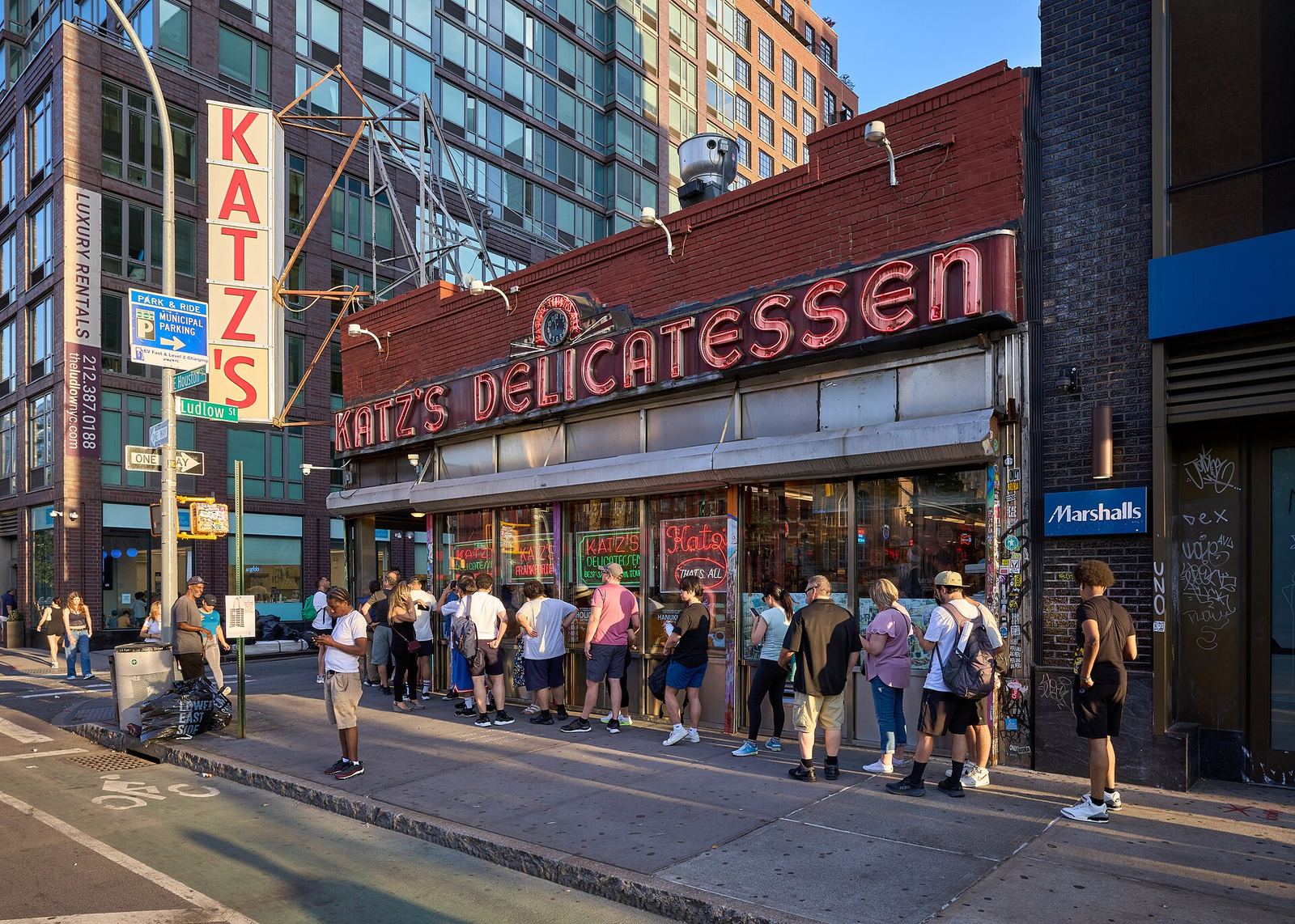 Katz's Delicatessen on the corner of Houston and Ludlow Streets, its red neon sign and tall vertical KATZ'S sign above a line of people waiting outside in the evening sun