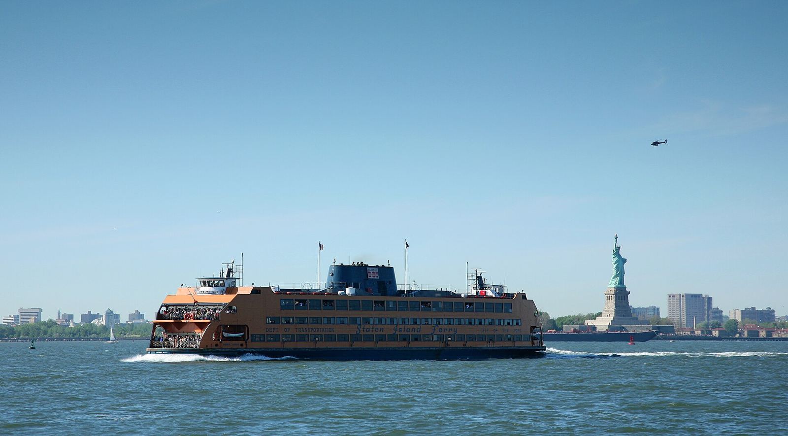 An orange Staten Island ferry, the Andrew J. Barberi, crossing New York Harbor under a clear blue sky, with the Statue of Liberty on its island behind it