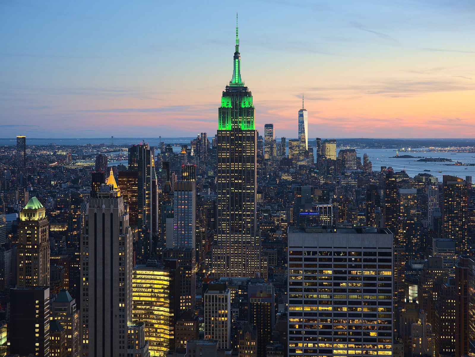 The Empire State Building lit green at dusk, seen from Top of the Rock above Midtown's lit-up towers, with a pink and orange sunset along the horizon