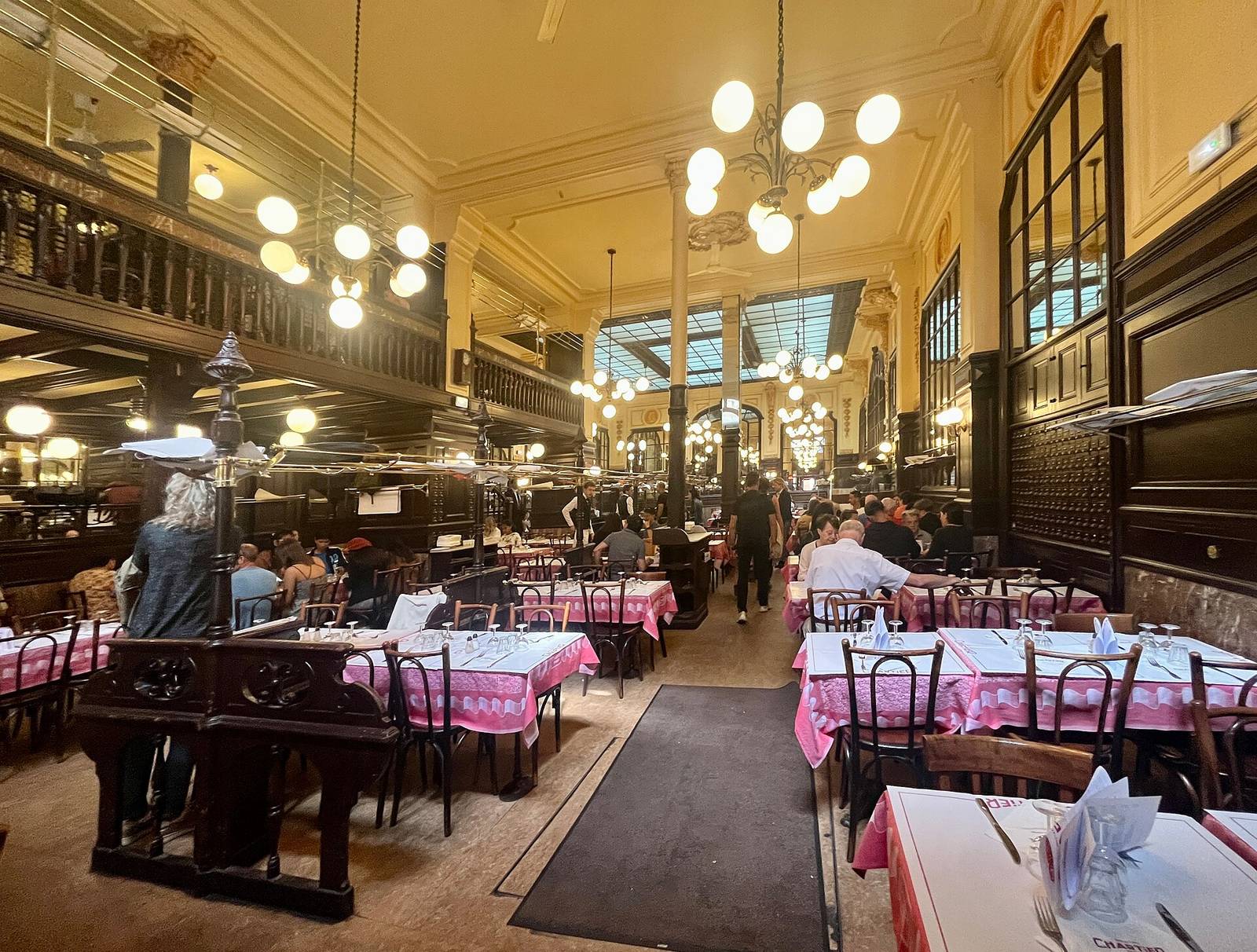 The long Belle Époque dining room of Bouillon Chartier, with globe lamps, a glass skylight, dark wood fittings and tables laid with pink cloths