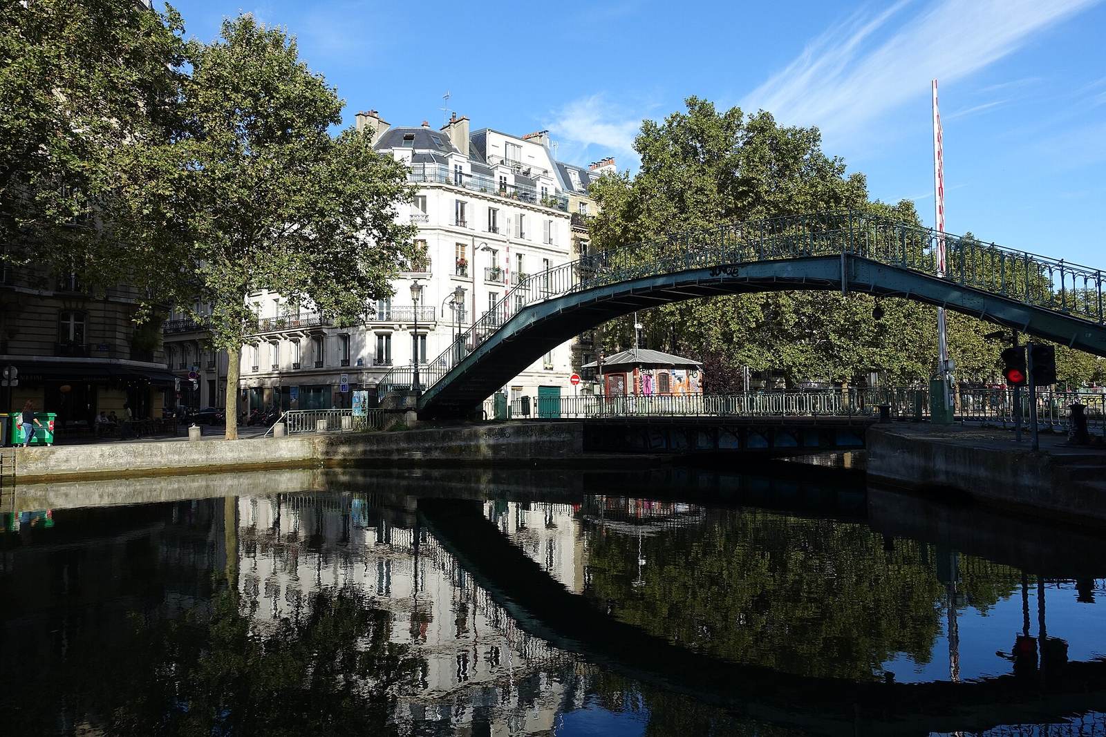 An arched iron footbridge over the Canal Saint-Martin, mirrored in the still water, with plane trees and a white Haussmann building behind