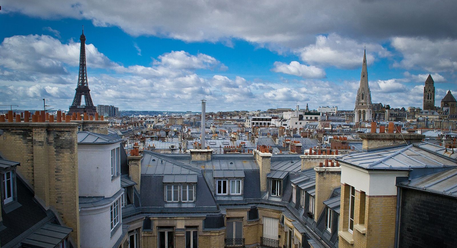 Grey zinc rooftops, chimney pots and dormer windows stretching across western Paris, with the Eiffel Tower on the left and a Gothic church spire on the right under blue sky and white clouds