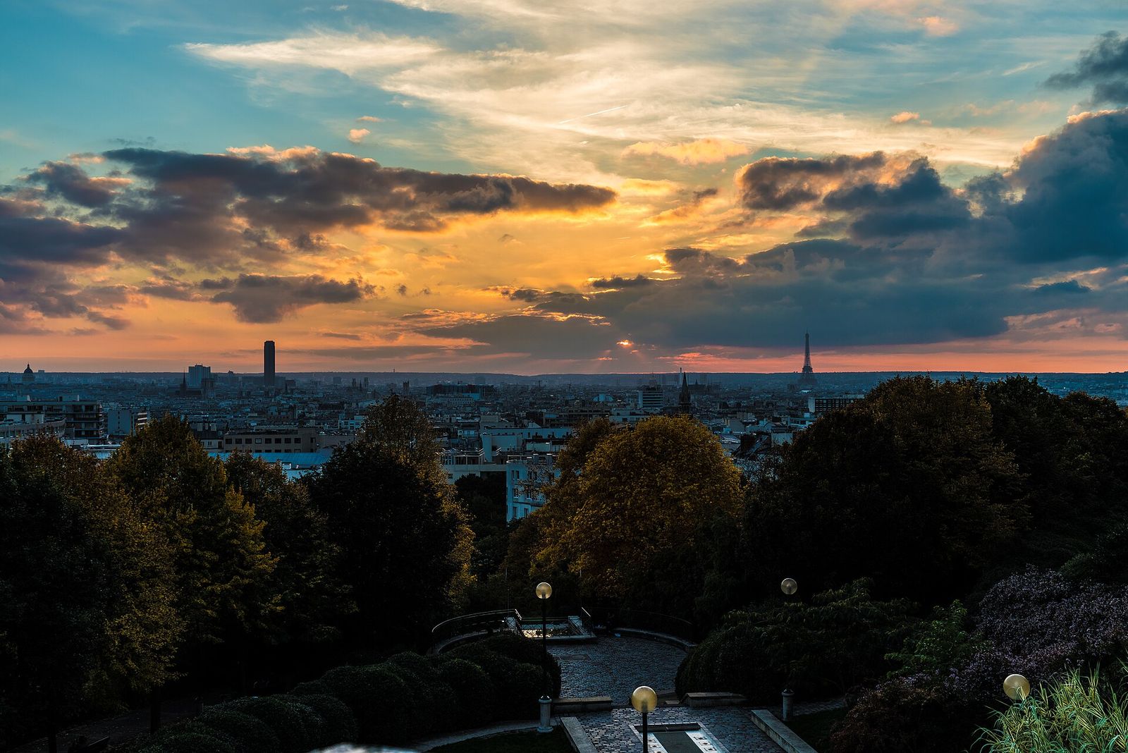 Sunset over Paris from the top of the Parc de Belleville, with the Eiffel Tower and the Tour Montparnasse silhouetted on the skyline above the park's trees and lamp-lit paths