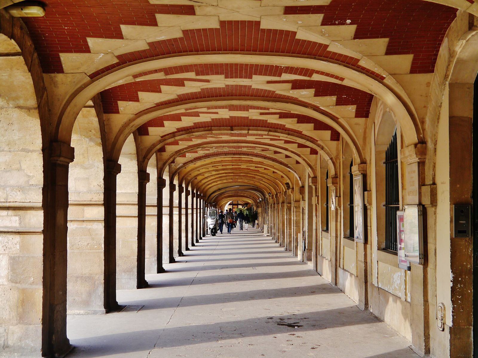 A sunlit arcade of the Place des Vosges: a long row of stone arches under red-brick and stone vaulting, receding into the distance
