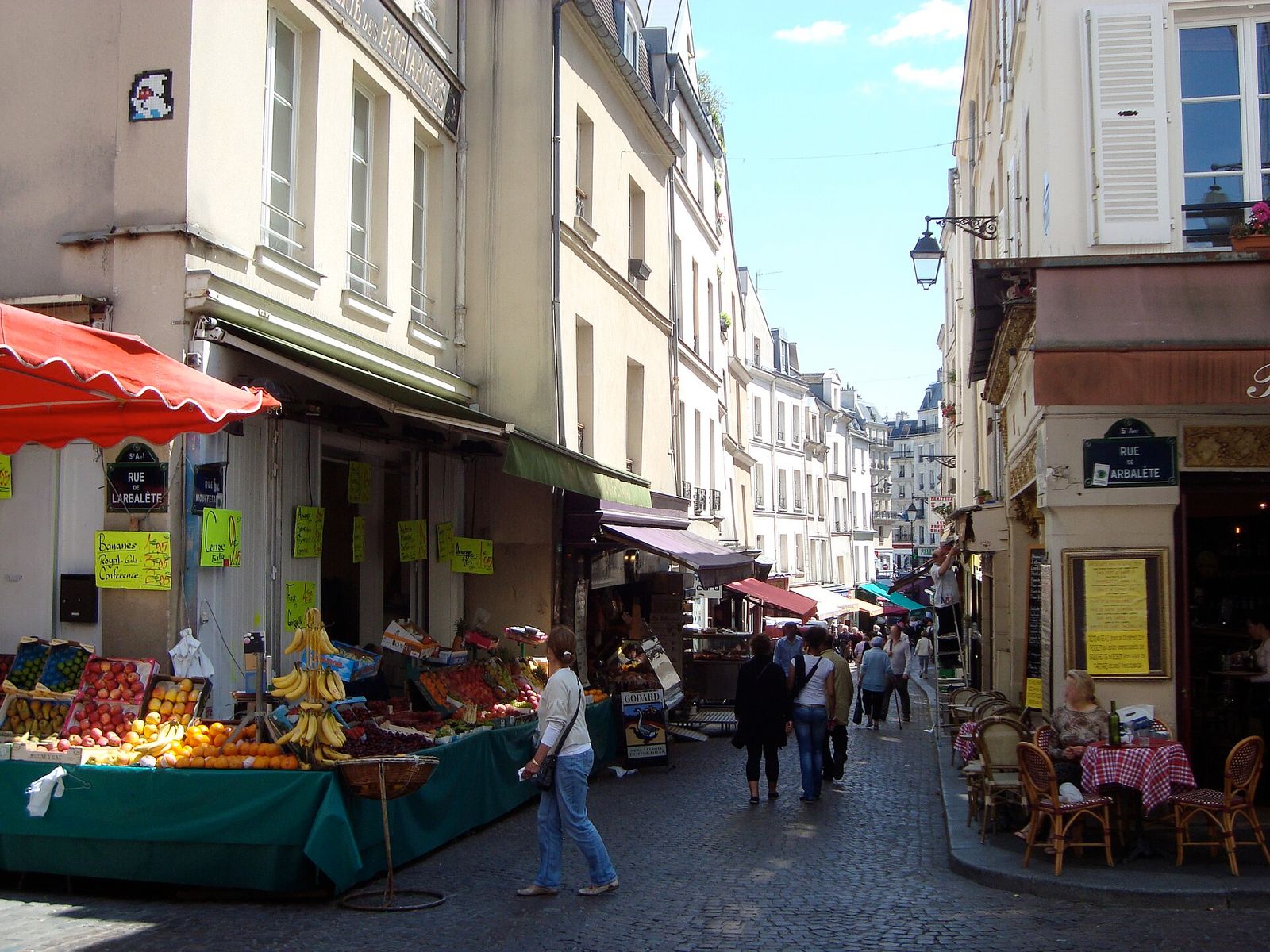 Fruit and vegetable stalls under red awnings on Rue Mouffetard at the corner of Rue de l'Arbalète, with shoppers walking down the narrow market street