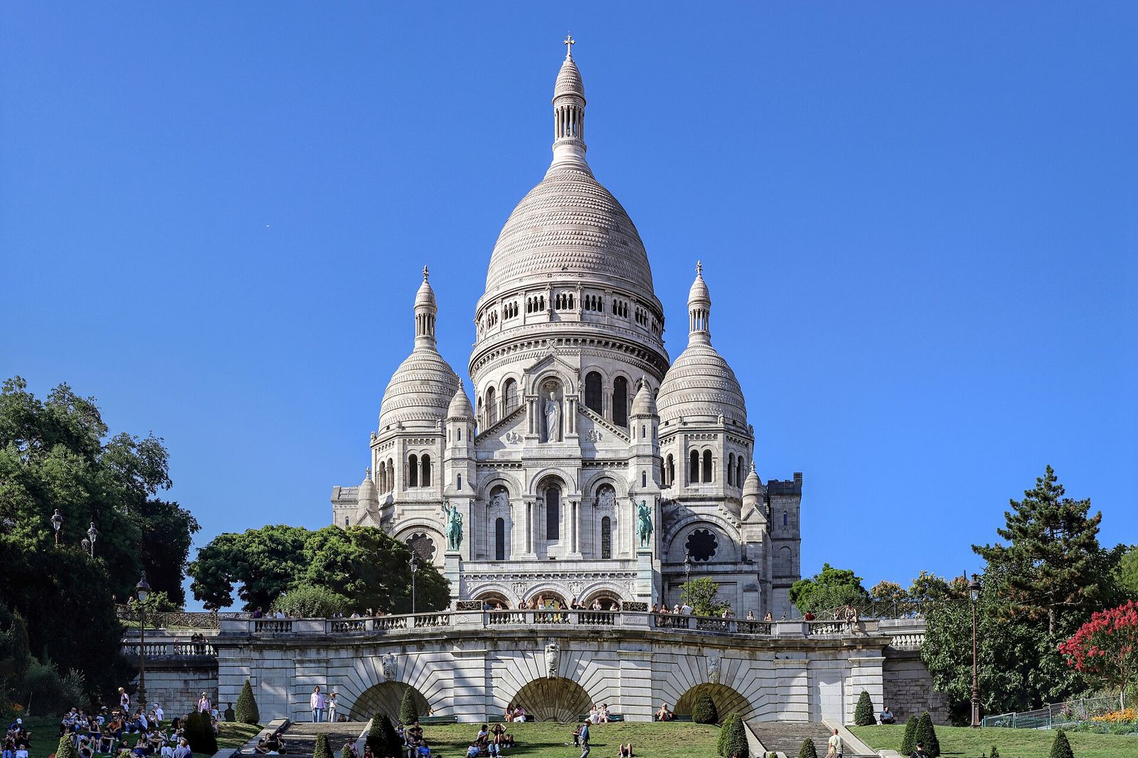 The white domes of Sacré-Cœur on top of Montmartre under a clear blue sky, above the terraced lawns and stairways of Square Louise-Michel where people sit in the sun