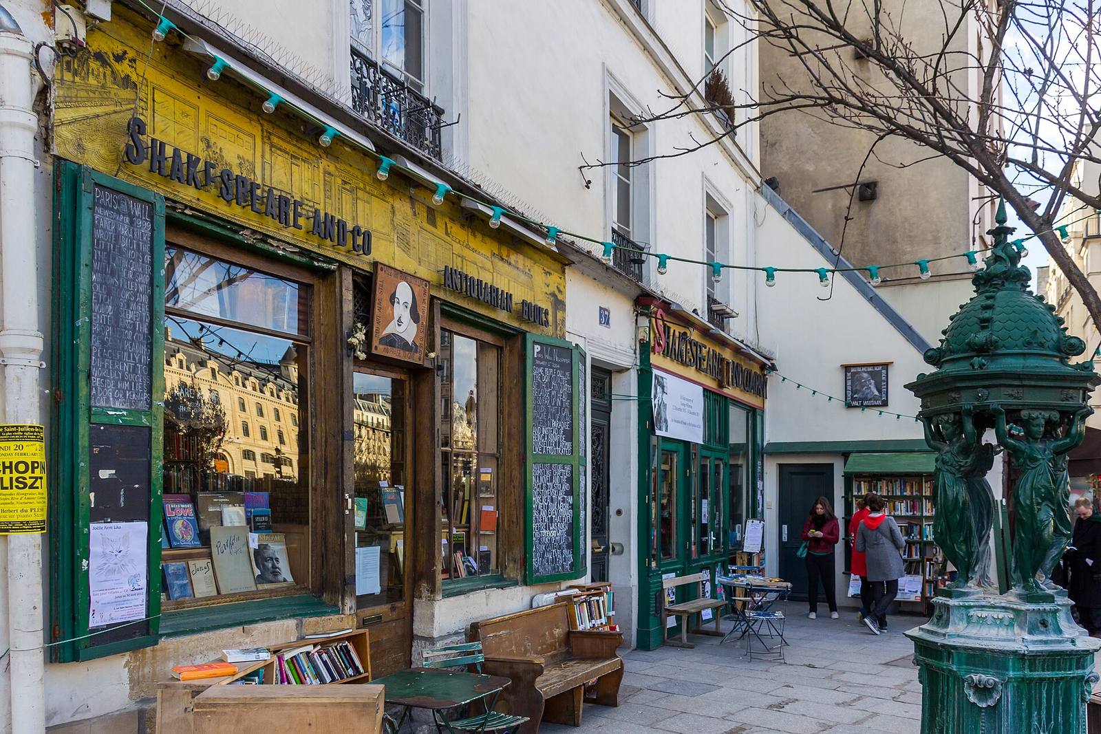 The green wooden shopfront of Shakespeare and Company under its yellow painted sign, books stacked in the windows, with a dark-green Wallace drinking fountain on the pavement to the right