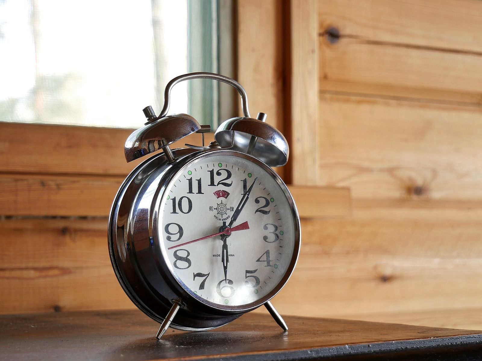 A chrome twin-bell mechanical alarm clock on a dark wooden shelf in front of pine-panelled walls and a window
