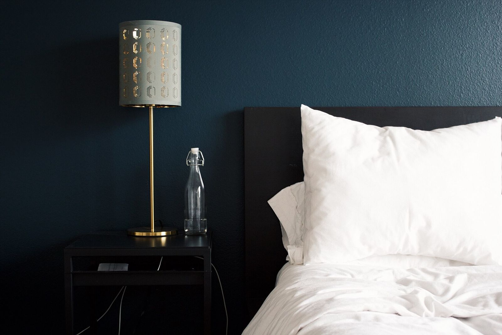 A black bedside table holding a brass lamp and a glass water bottle, next to a bed with white pillows against a dark teal wall