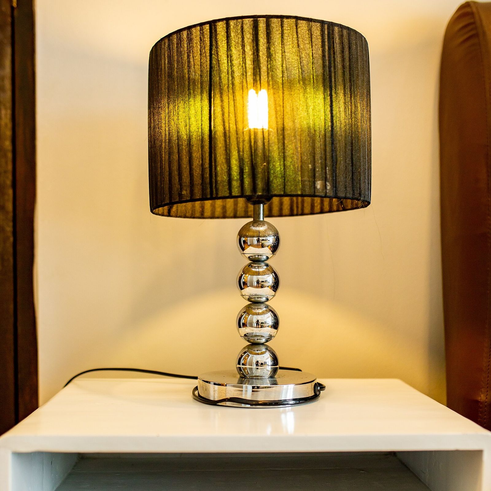 A lit table lamp with a dark pleated shade and a chrome base on a white bedside table, casting warm light on a cream wall