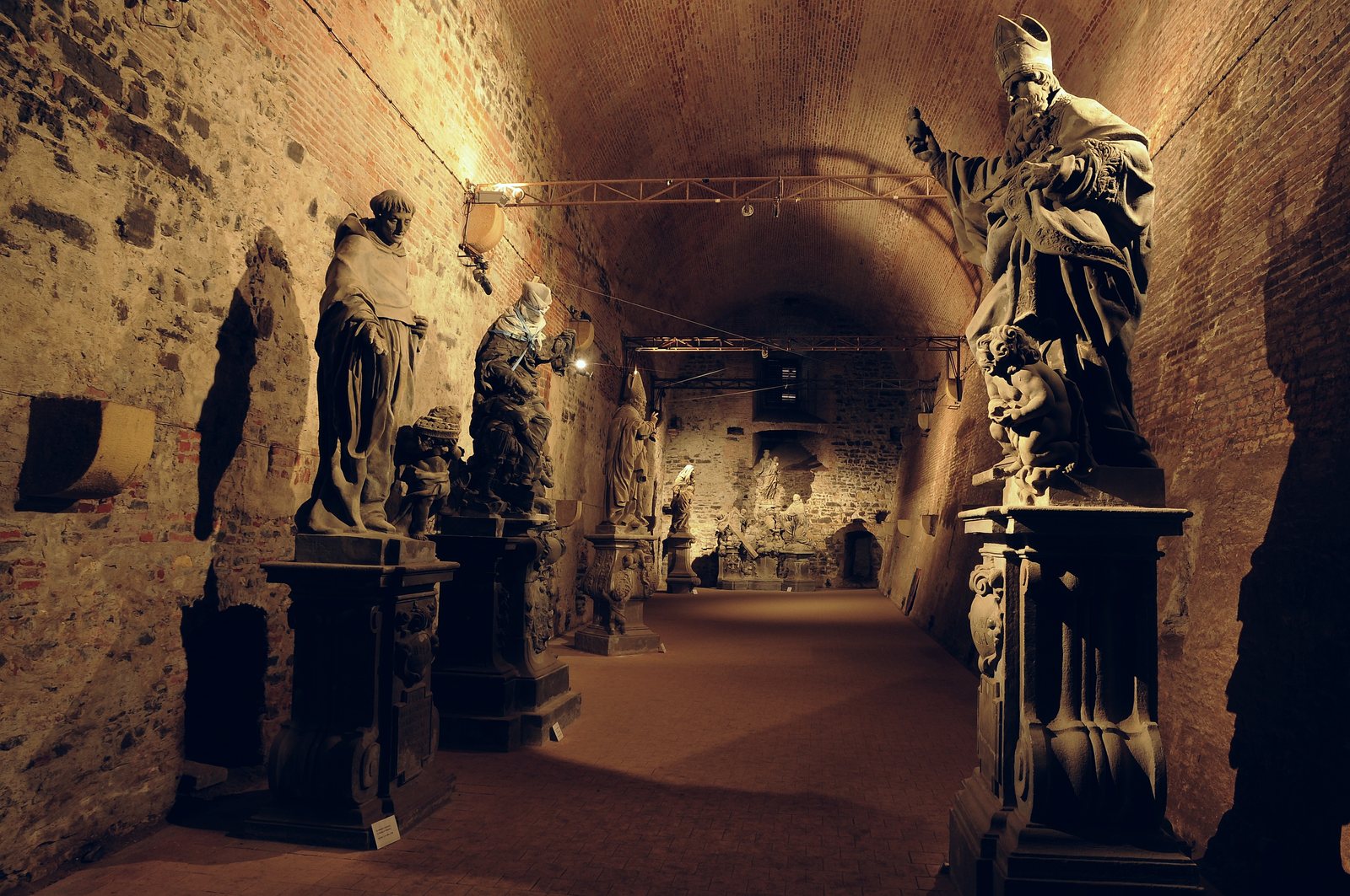Baroque stone saints from Charles Bridge on pedestals along a lit brick vault in the Gorlice hall under Vyšehrad