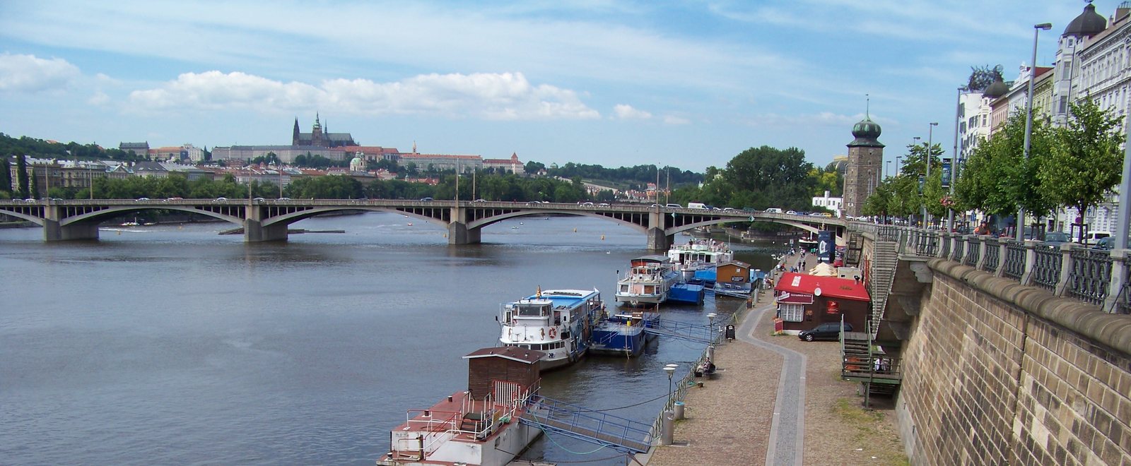 The Náplavka embankment on a summer day: river boats moored along the stone quay, Jirásek Bridge across the Vltava and Prague Castle on the skyline beyond