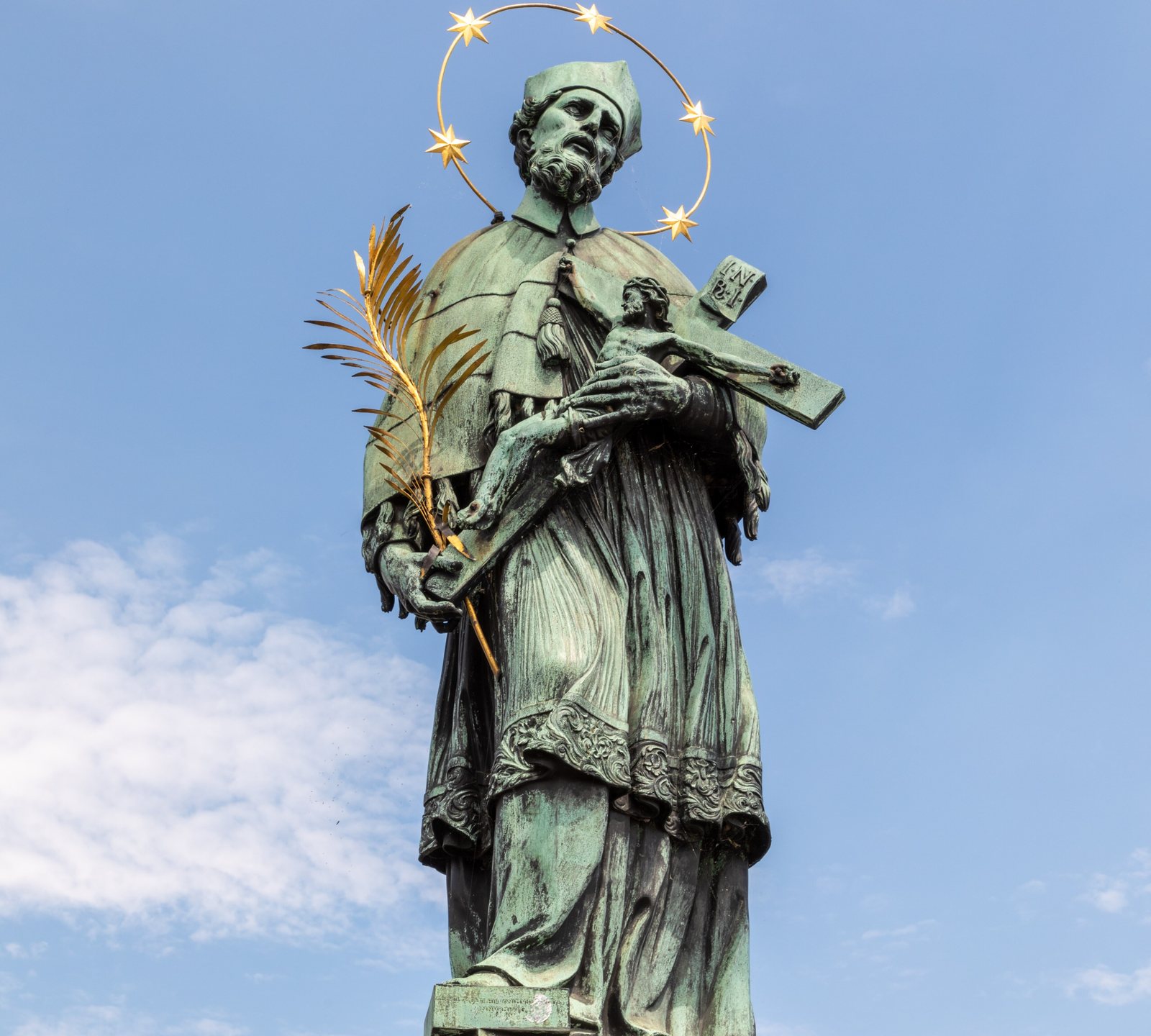 The bronze statue of St John of Nepomuk on Charles Bridge, holding a crucifix and a palm frond under a halo of five gold stars, against a blue sky
