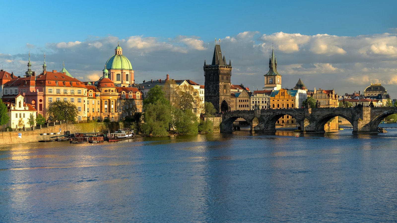 The Old Town Bridge Tower and the first arches of Charles Bridge in golden evening light, with the green dome of St Francis of Assisi and the Old Town's riverside houses beside it, seen across the Vltava