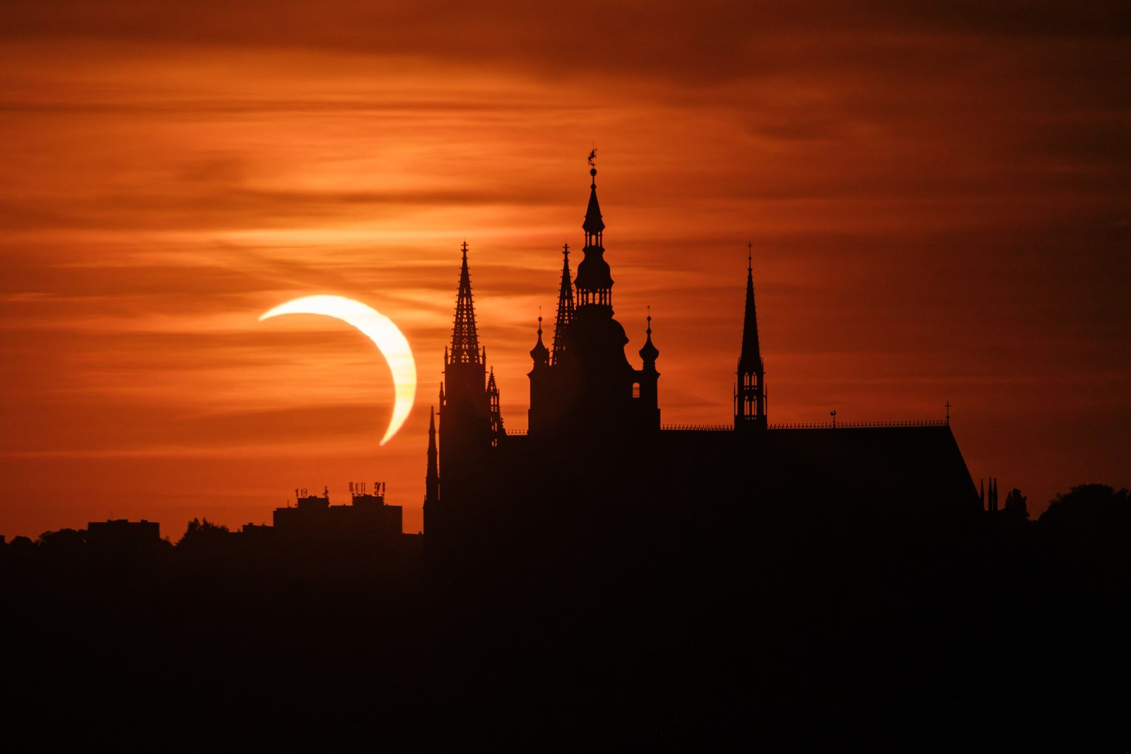 Prague Castle and St Vitus Cathedral in silhouette against an orange sky, with a crescent sun during the partial solar eclipse of 12 August 2026, seen from Riegrovy sady