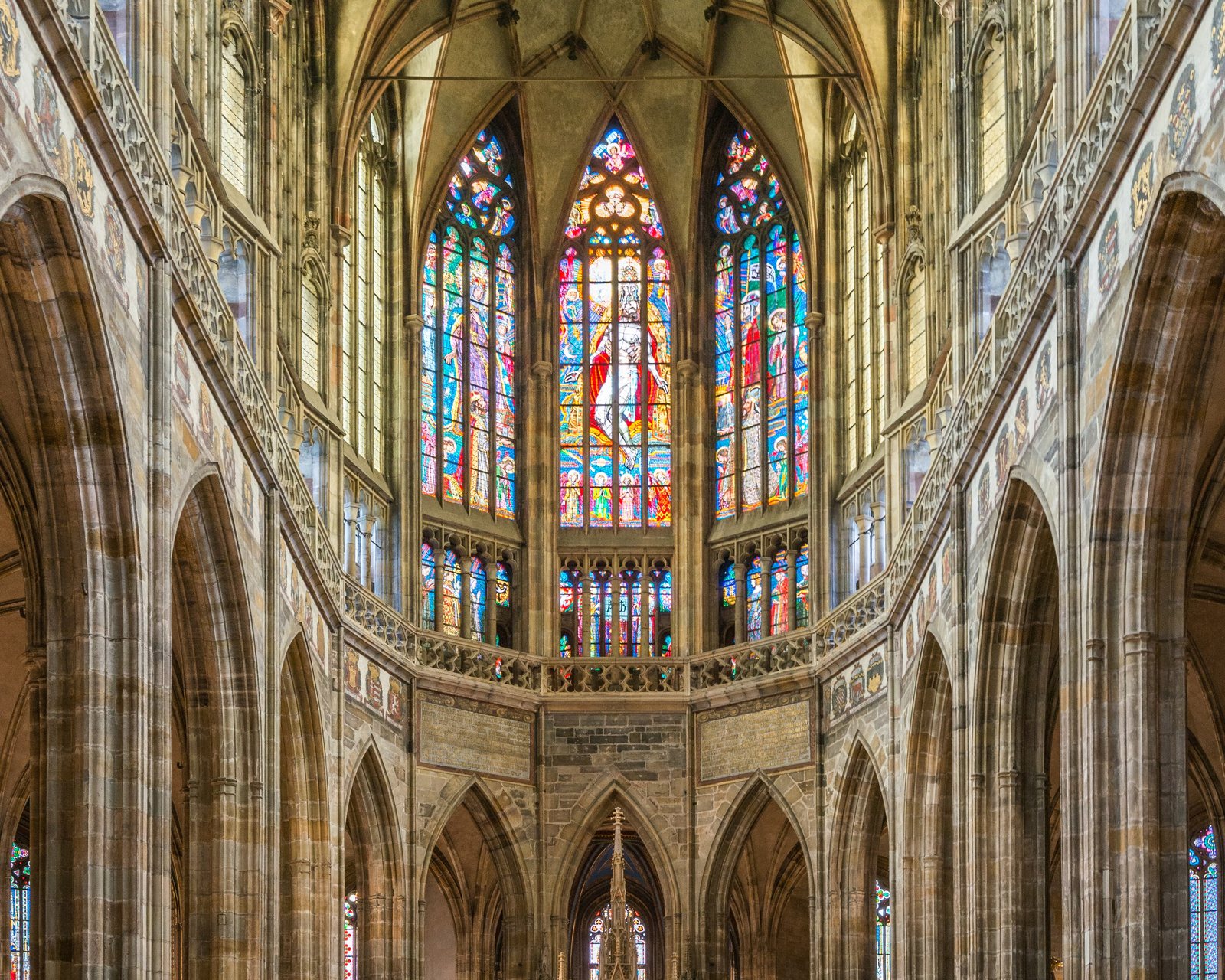 The choir of St Vitus Cathedral: Gothic vaulting and tall columns around three stained-glass windows, above the high altar and a wrought-iron screen