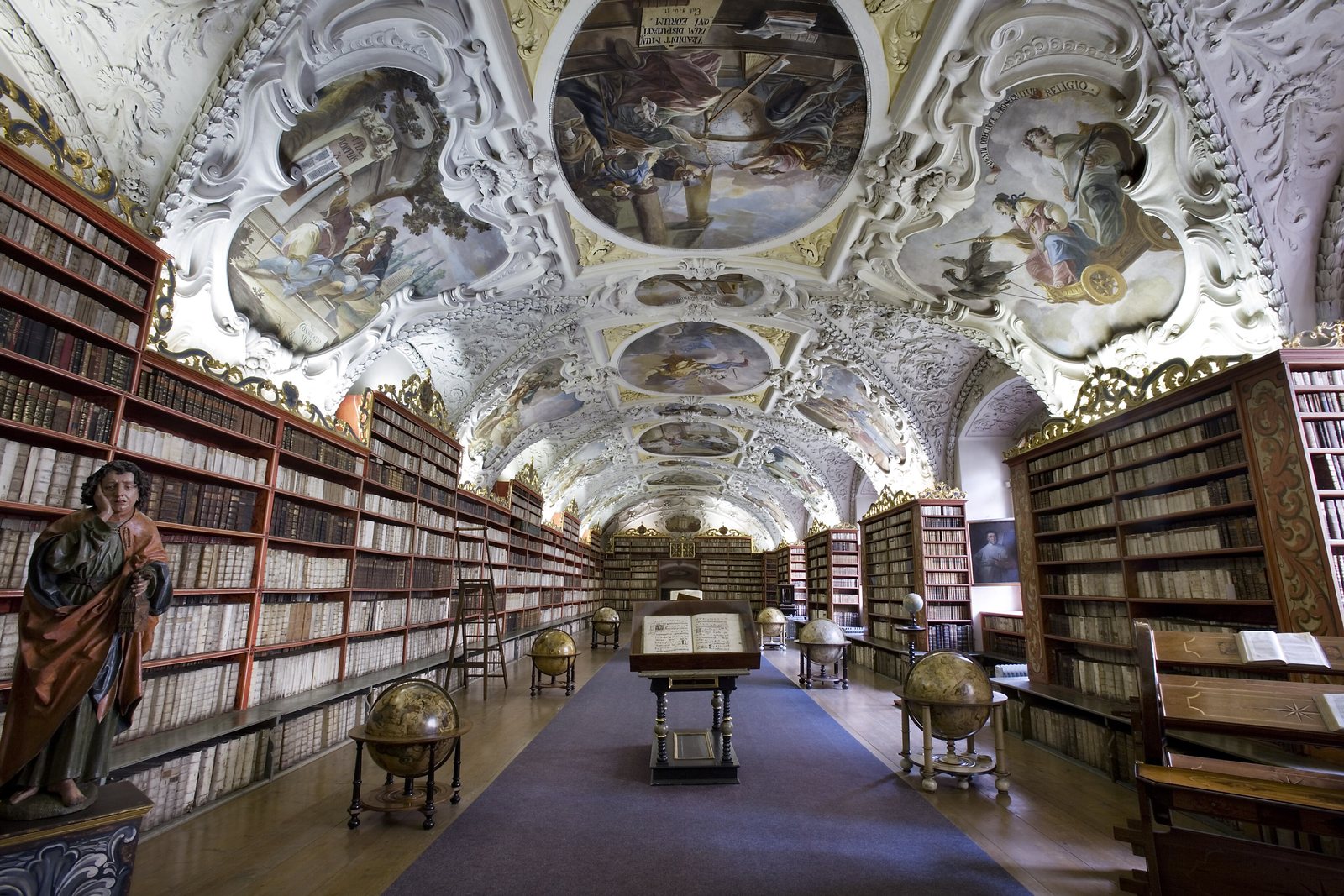 The Theological Hall of the Strahov Library: gilded Baroque bookcases along both walls under a white stucco vault painted with frescoes, with antique globes down the middle