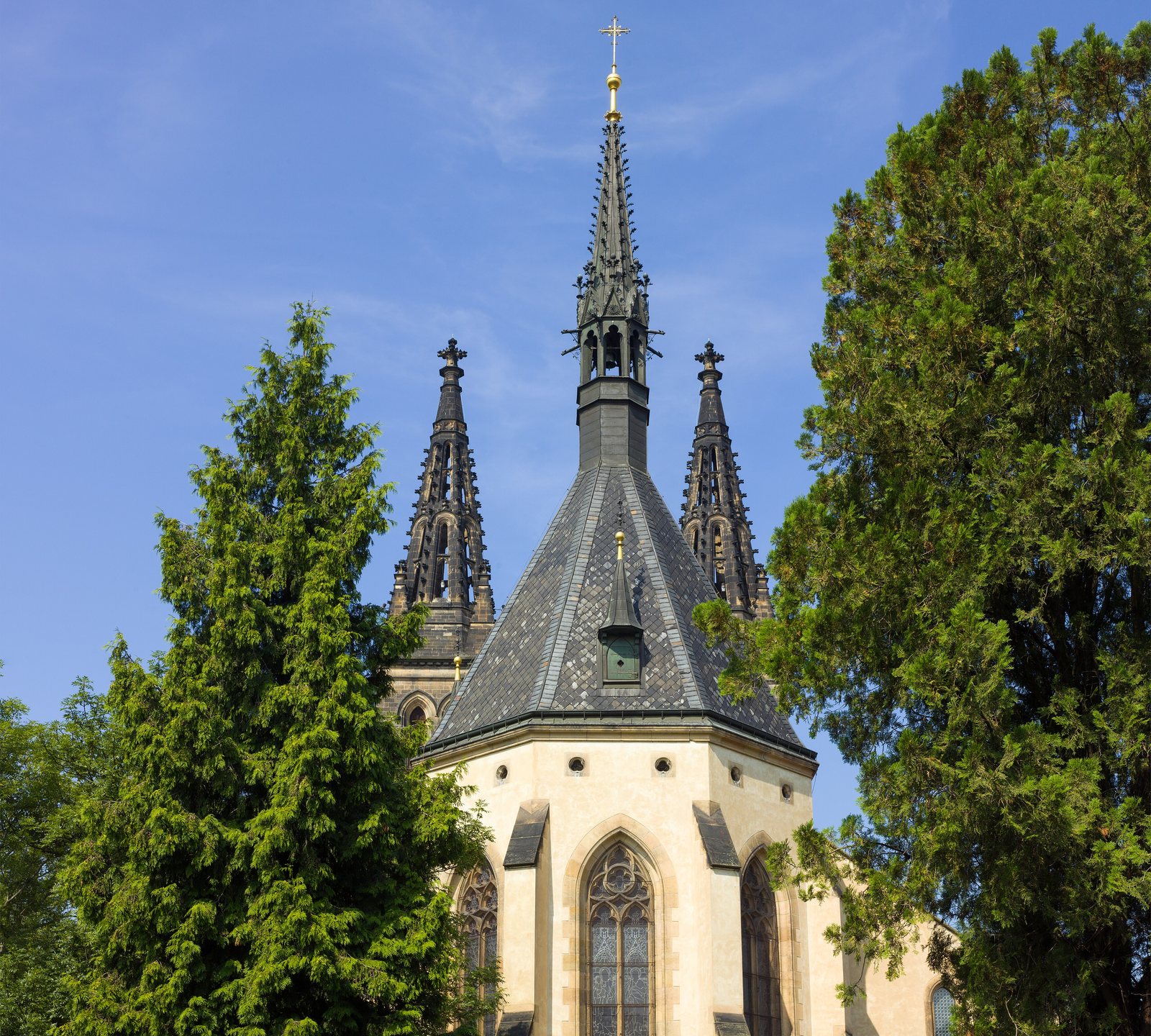 The Basilica of Sts Peter and Paul at Vyšehrad seen from the cemetery: a pale stone apse under a steep slate roof and three dark spires, between tall trees