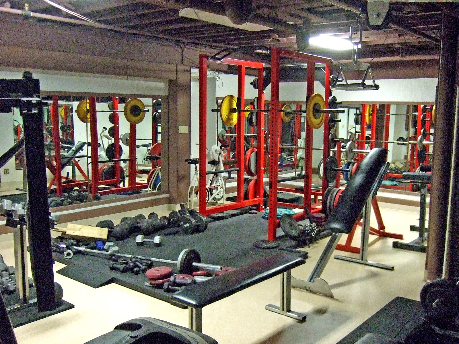 An empty basement weights room with two red power racks holding barbells loaded with yellow plates, an incline bench in the foreground, and dumbbells and plates on a black rubber floor, all reflected in a mirrored wall