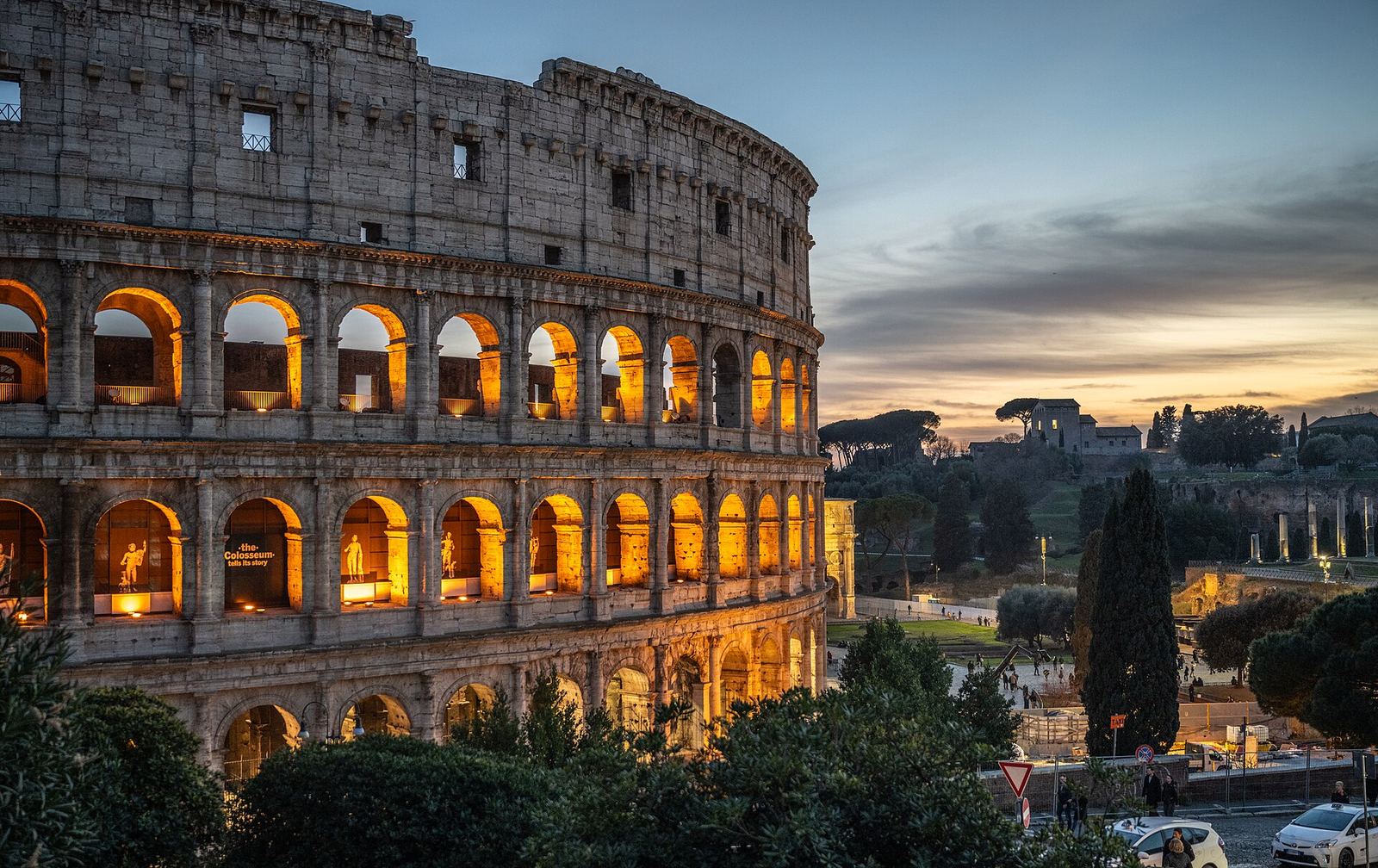 The Colosseum at dusk, its lower arcades lit gold, with umbrella pines and the edge of the Palatine Hill under a pink and blue sky