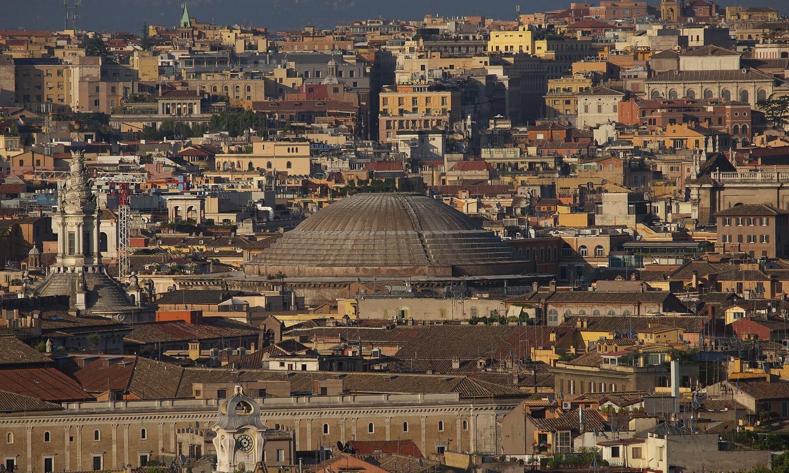 The Pantheon's grey dome rising among the ochre and terracotta rooftops of Rome's historic center, with the white spiral lantern of Sant'Ivo alla Sapienza to its left, seen from the Gianicolo hill in warm late-afternoon light
