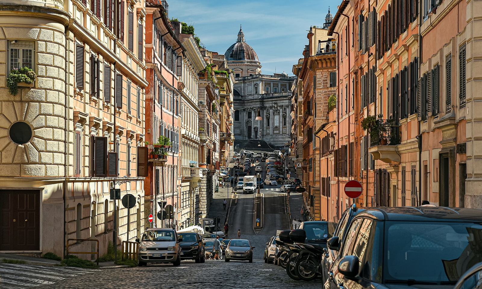 Via Panisperna in Monti, the street Ai Tre Scalini is on, lined with ochre and pink buildings and parked cars and running toward the dome of Santa Maria Maggiore