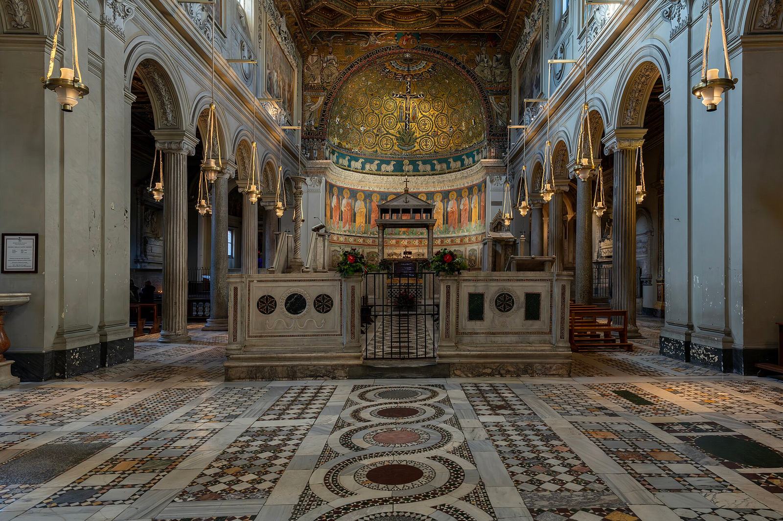 The nave of San Clemente: a patterned marble floor leads to the walled choir and a gold apse mosaic with a cross at its center, under hanging lamps