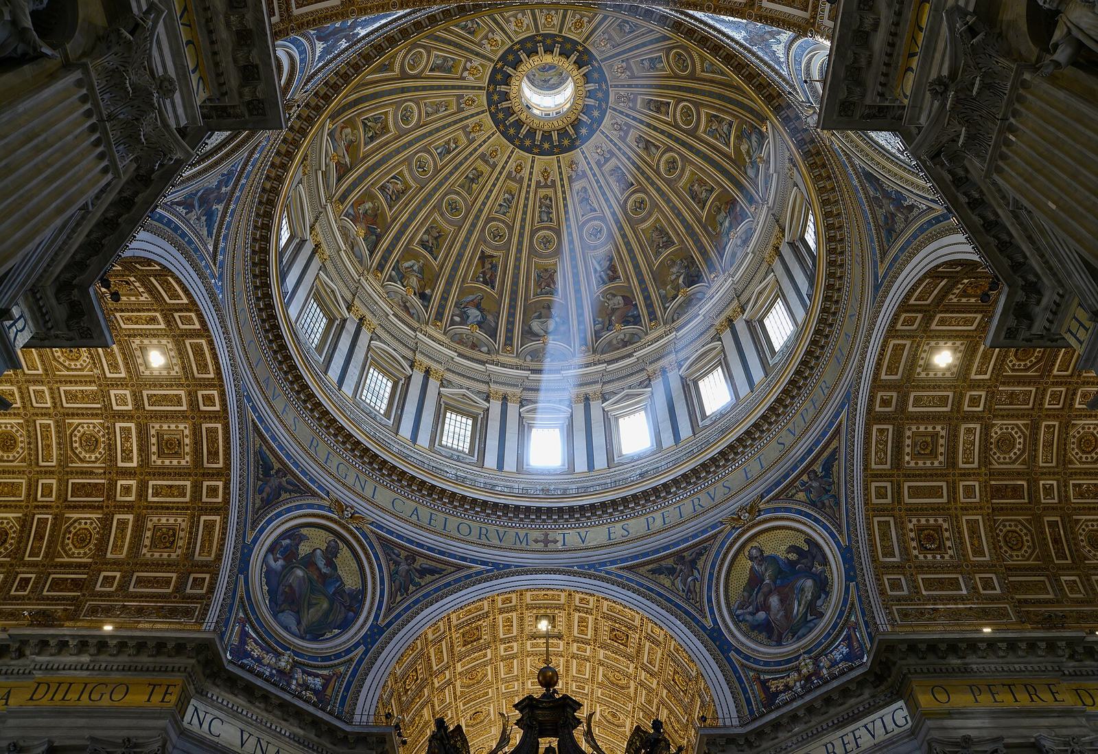 Looking straight up into the dome of St Peter's: gold coffering, a ring of windows letting in beams of sunlight and the Latin inscription around its base, with the top of Bernini's canopy at the bottom